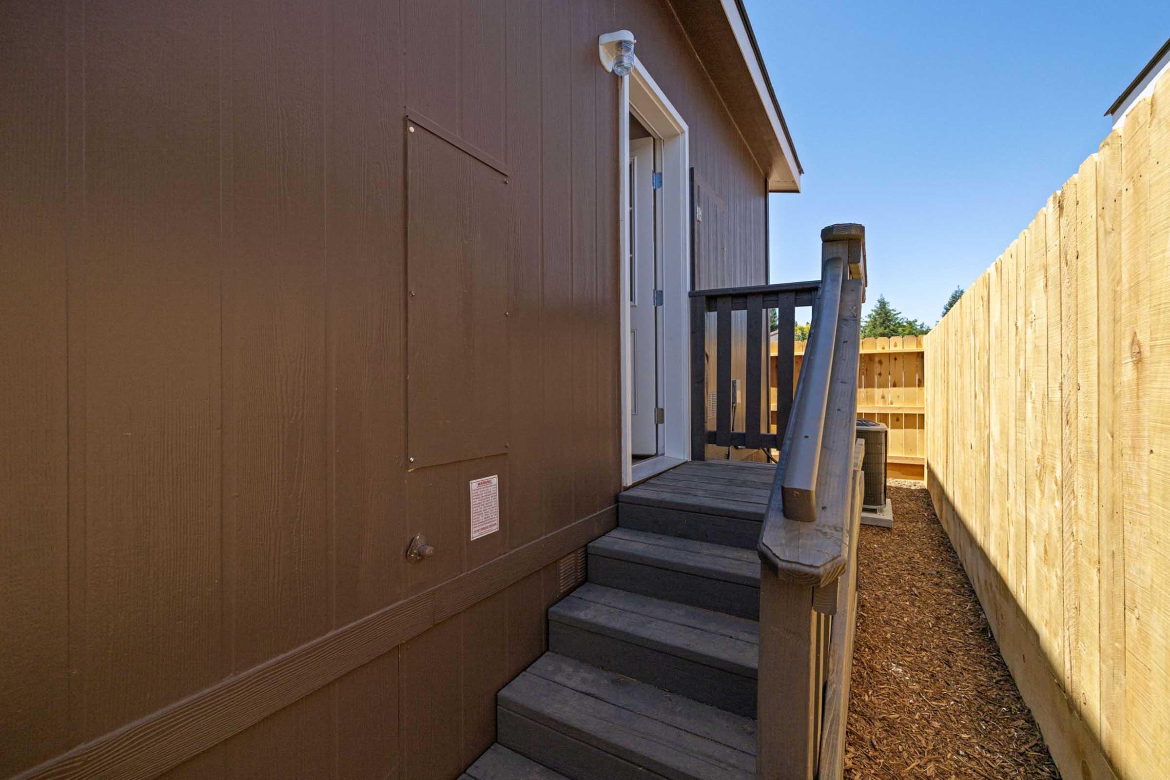 A view of a brown wooden building with a set of stairs leading up to a door. There is a fenced area on the right, and the ground is covered with mulch. A small wall-mounted fixture is visible next to the door. The sky is clear and blue.