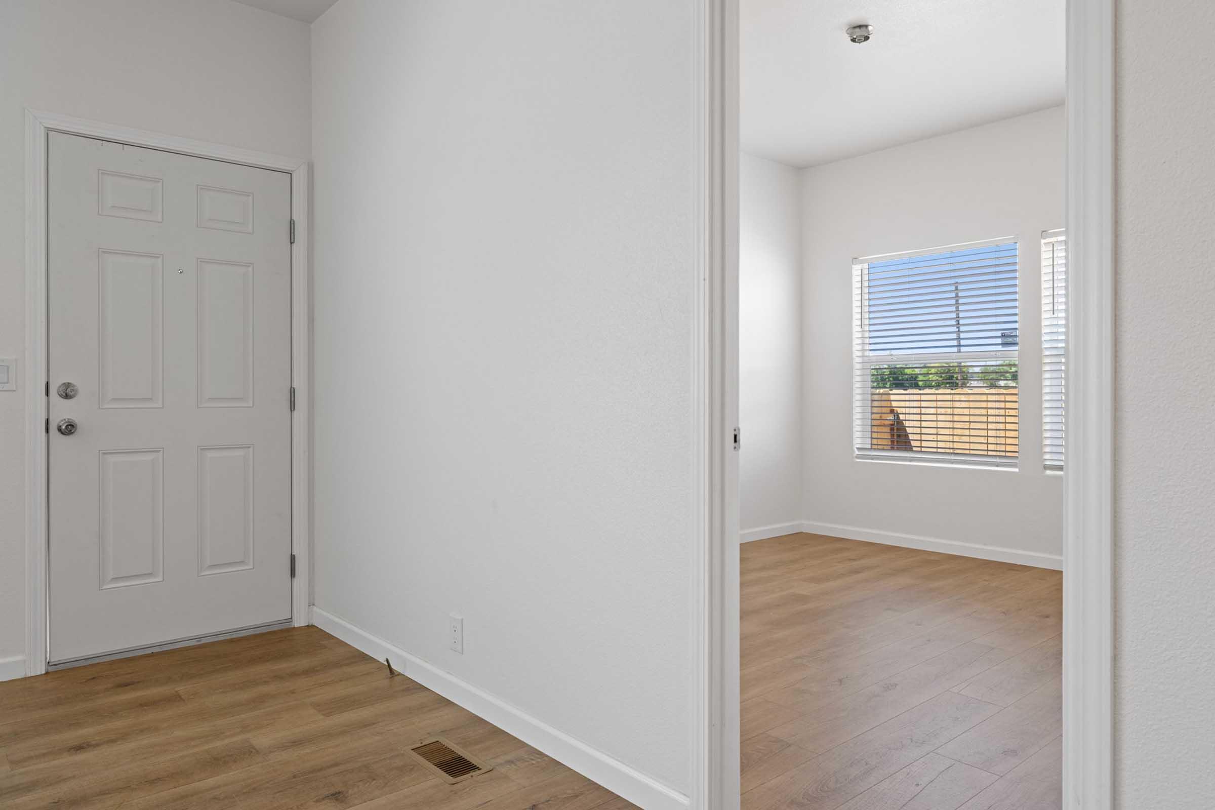 A view from a hallway into a well-lit room with a window. The walls are painted white, and the flooring is light wood. The front door is visible on the left with a simple design, and the room beyond has no furniture, highlighting its spaciousness and potential for customization.