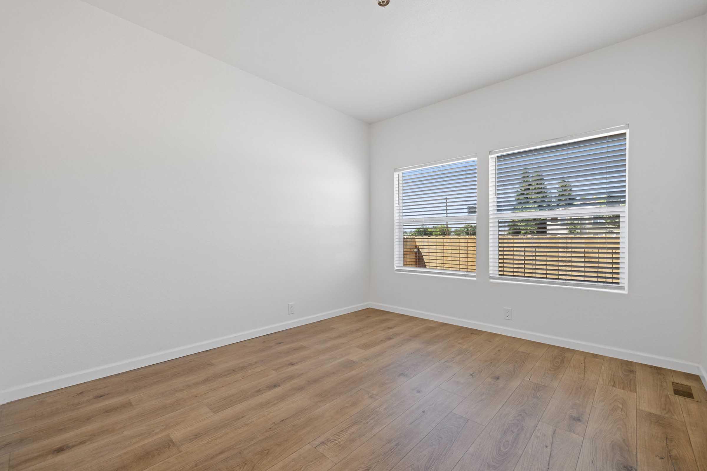 Empty room with light-colored walls and wooden flooring. Two windows with white blinds allow natural light to enter, illuminating the space. The room is unfurnished and has a clean, minimalist appearance, ideal for various decorating options.