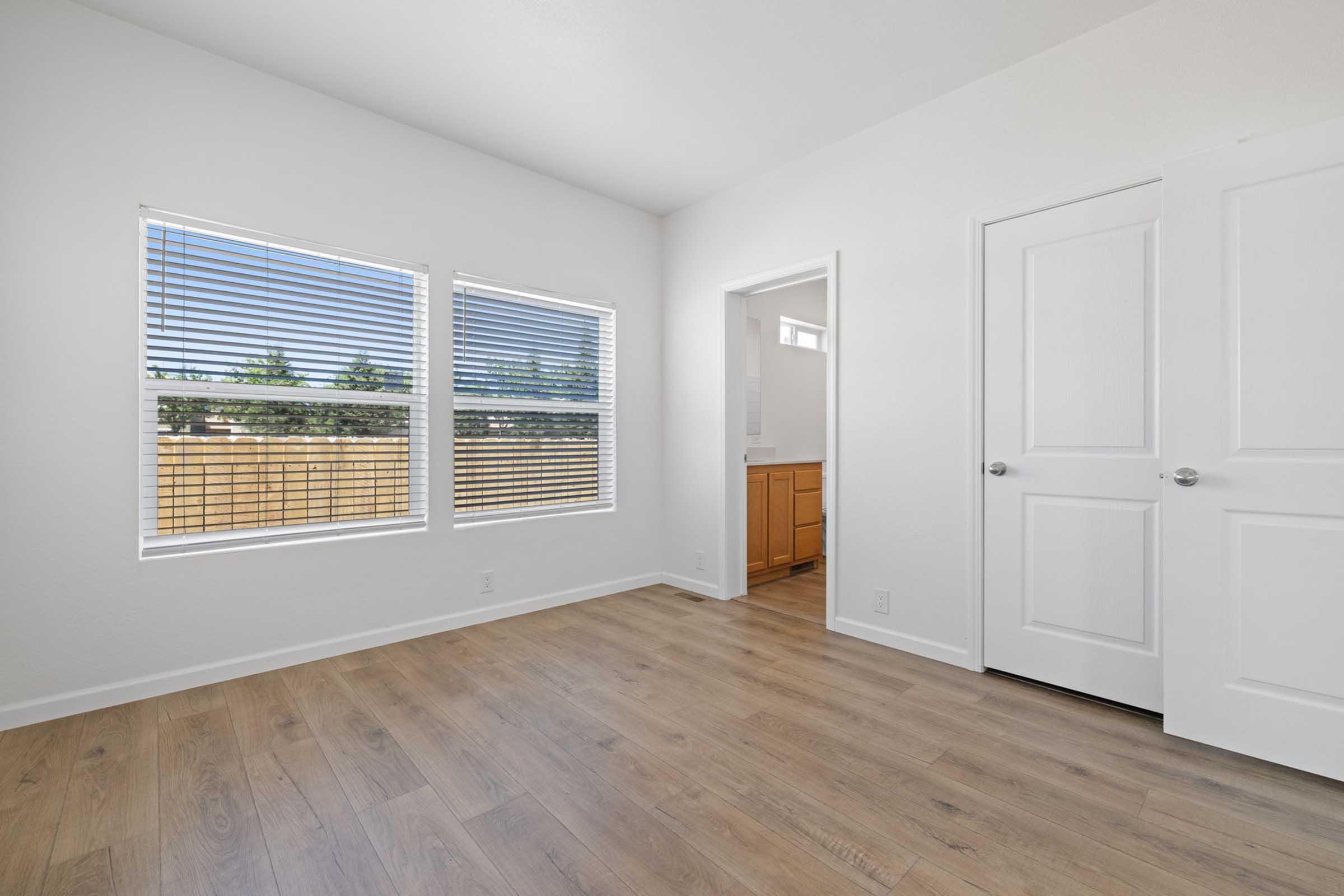 Empty room with light-colored walls and hardwood flooring. There are two large windows with white blinds allowing natural light, and a doorway leading to an adjacent area. The space has a minimalistic design and appears well-lit and inviting.