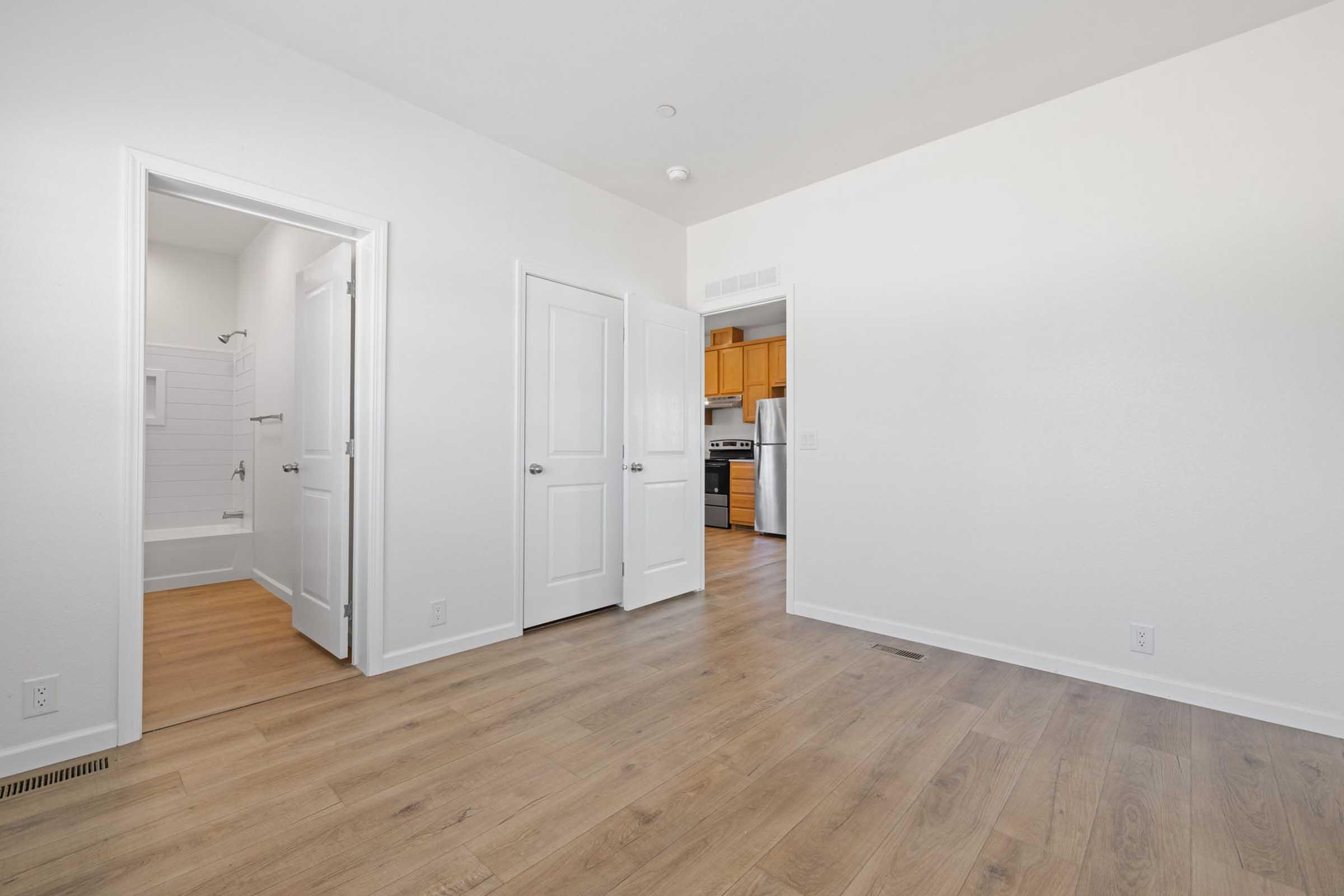 A spacious interior room featuring light-colored walls and wood laminate flooring. In the foreground, a doorway leads to another room and a bathroom. The background shows an open kitchen area with wooden cabinets. Natural light brightens the room, creating an inviting atmosphere.