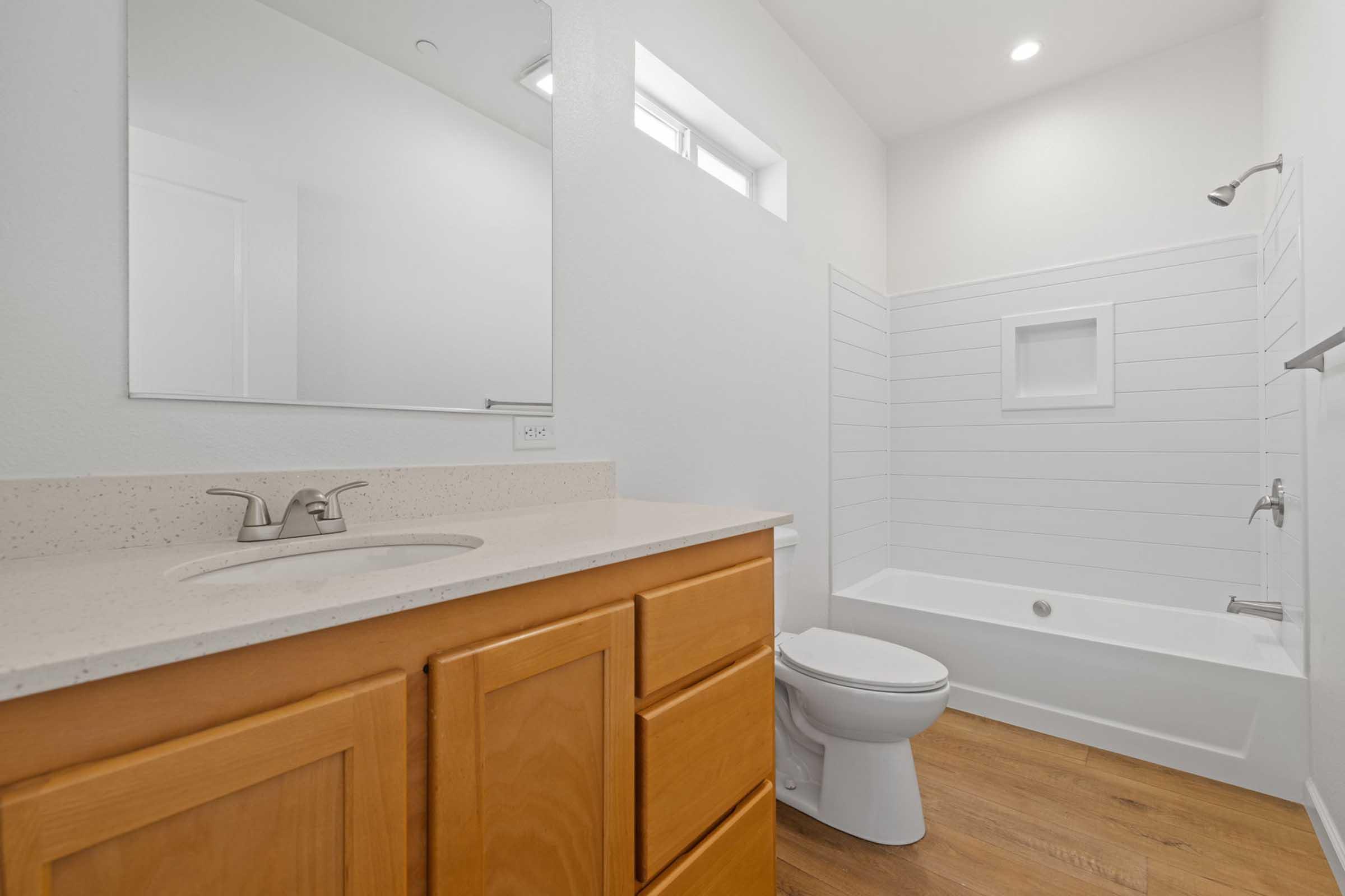 A modern bathroom featuring a wooden vanity with a sink, a white toilet, and a shower area with white shiplap walls. The space is well-lit with natural light coming from a small window, and there is a large mirror above the sink reflecting the minimalist design.