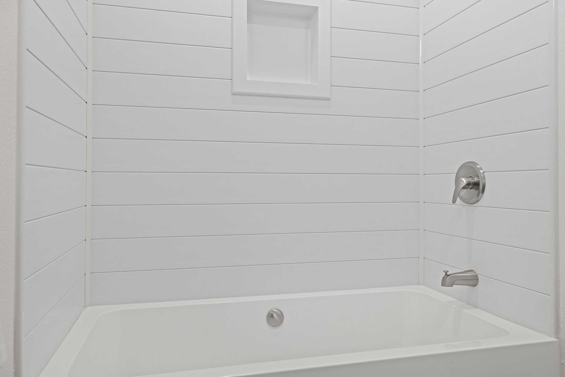 A modern, minimalistic bathroom featuring a white tub against a paneled wall. The shower fixture is mounted on the wall, and there is a small shelf built into the wall above the tub, contributing to a clean and contemporary aesthetic.