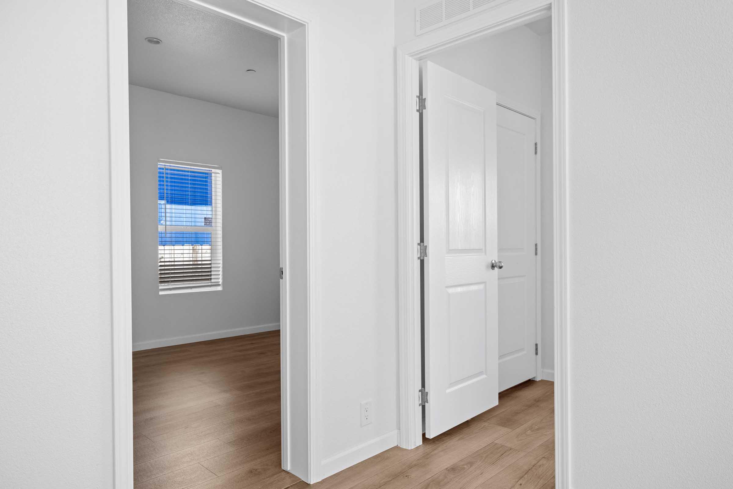 A view of a hallway with two doorways leading to different rooms. One doorway is partially open, revealing a room with white walls and a window. The other door is closed, and the floor features light-colored wooden flooring. Natural light brightens the area, enhancing the spacious feel.