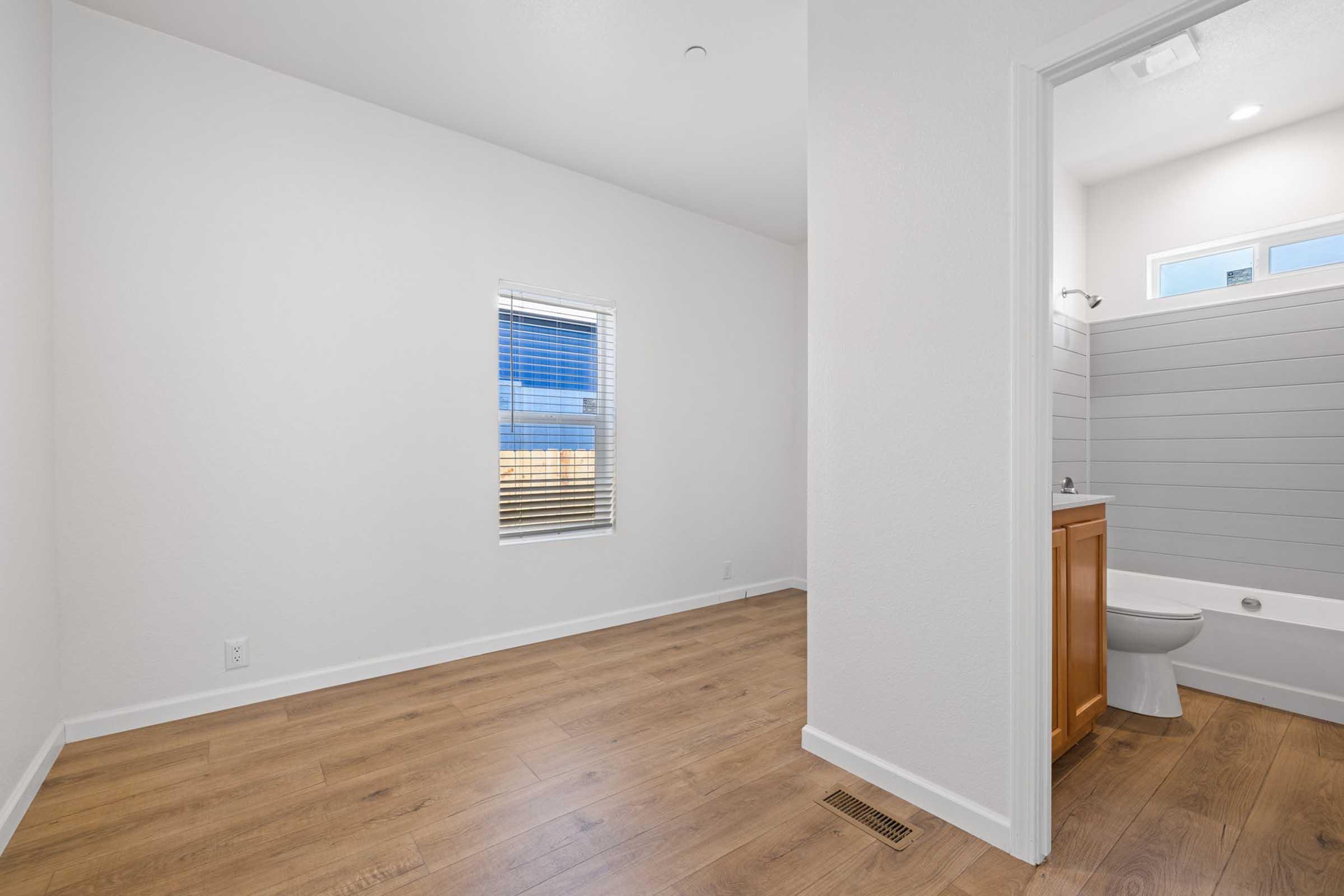 Empty room with light-colored walls, wooden flooring, and a window allowing natural light. A bathroom with a wooden vanity and a toilet is visible through an open doorway. The overall space appears modern and bright, with minimal decor.