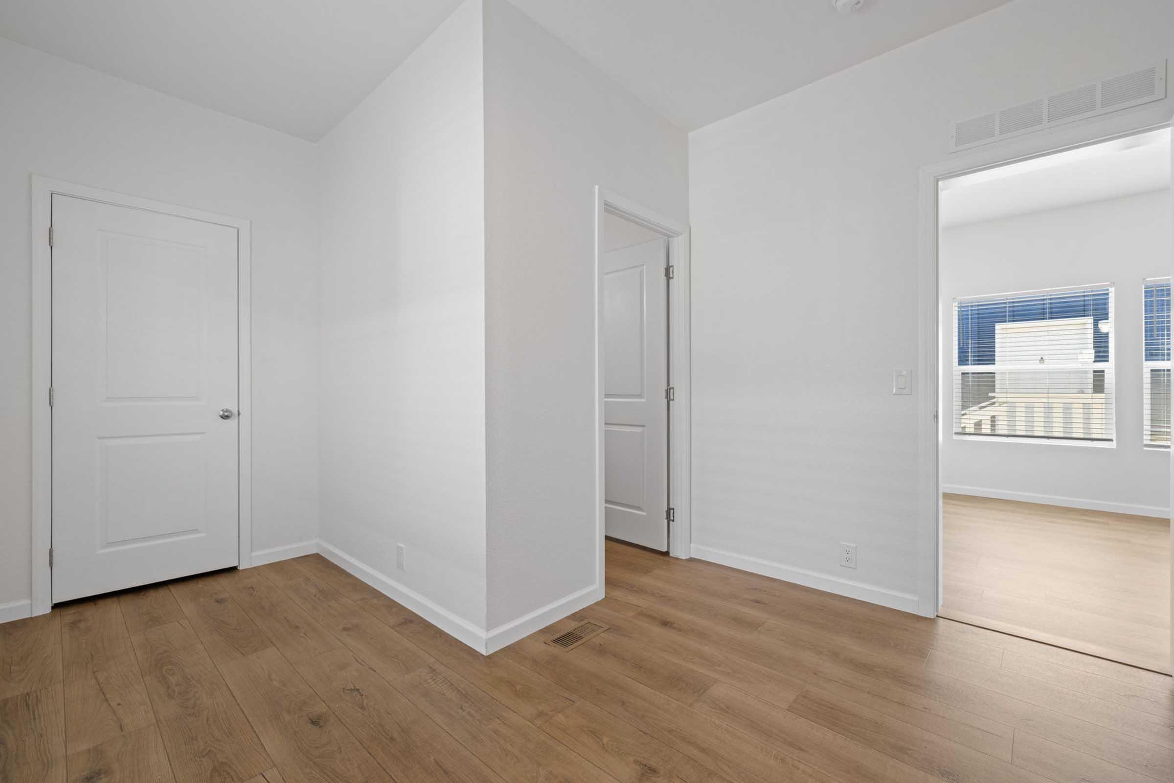 A well-lit, empty room featuring light-colored walls and wooden flooring. A doorway on the left leads to another room, while a window on the right provides natural light. The space is clean and modern, with minimal furnishings, creating an open atmosphere suitable for various uses.