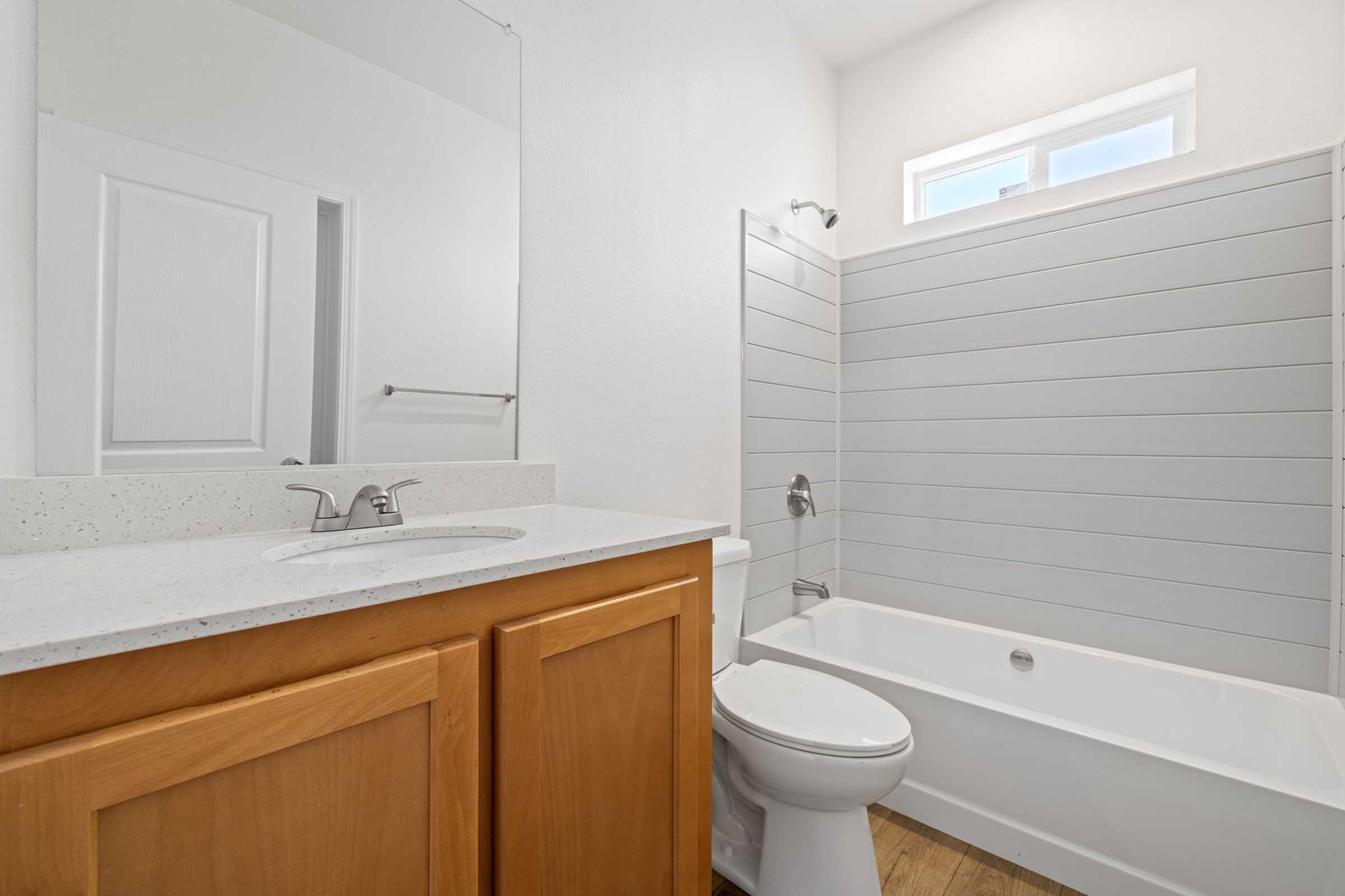 A modern bathroom featuring a white bathtub, a glass shower, a light wood cabinet with a sink, and a mirror. The walls are painted white with a shiplap design in the shower area. Natural light enters through a window above the tub, highlighting the clean and minimalistic design.