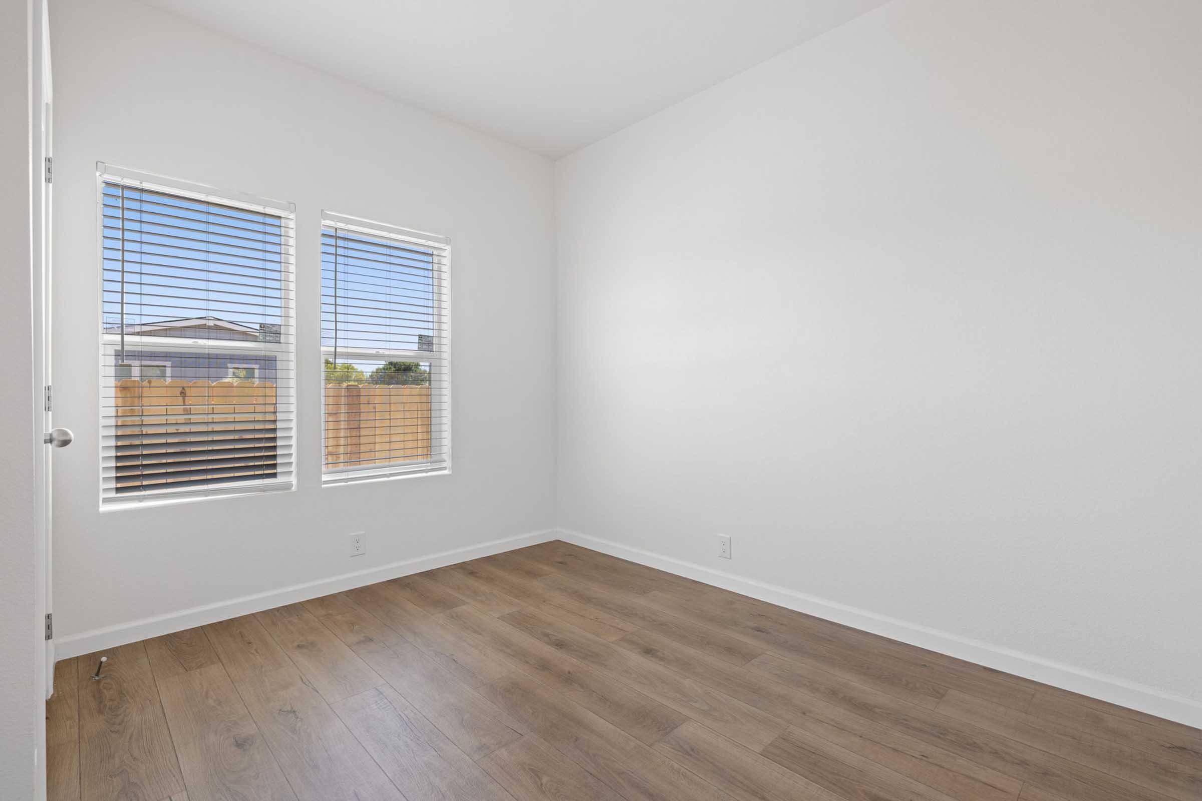 Empty room with light-colored walls, two windows with horizontal blinds allowing natural light, and wooden flooring. The view outside includes a wooden fence. The space feels bright and open, suitable for various purposes.