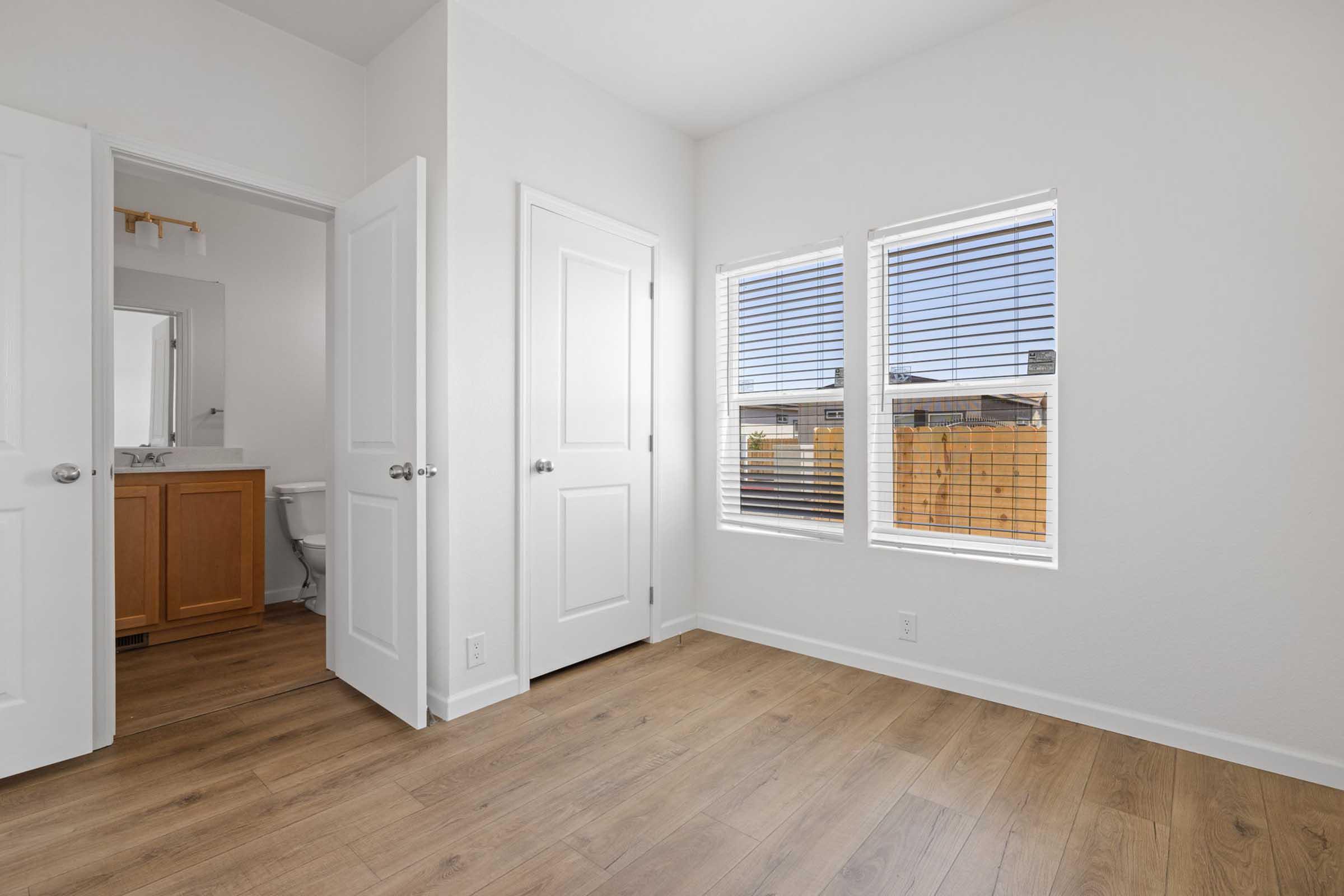 Bright, empty room featuring light-colored walls and wooden flooring. Two windows allow natural light, with white blinds partially closed. A doorway leads to a bathroom, visible in the background, and another door is slightly open, revealing more of the space.