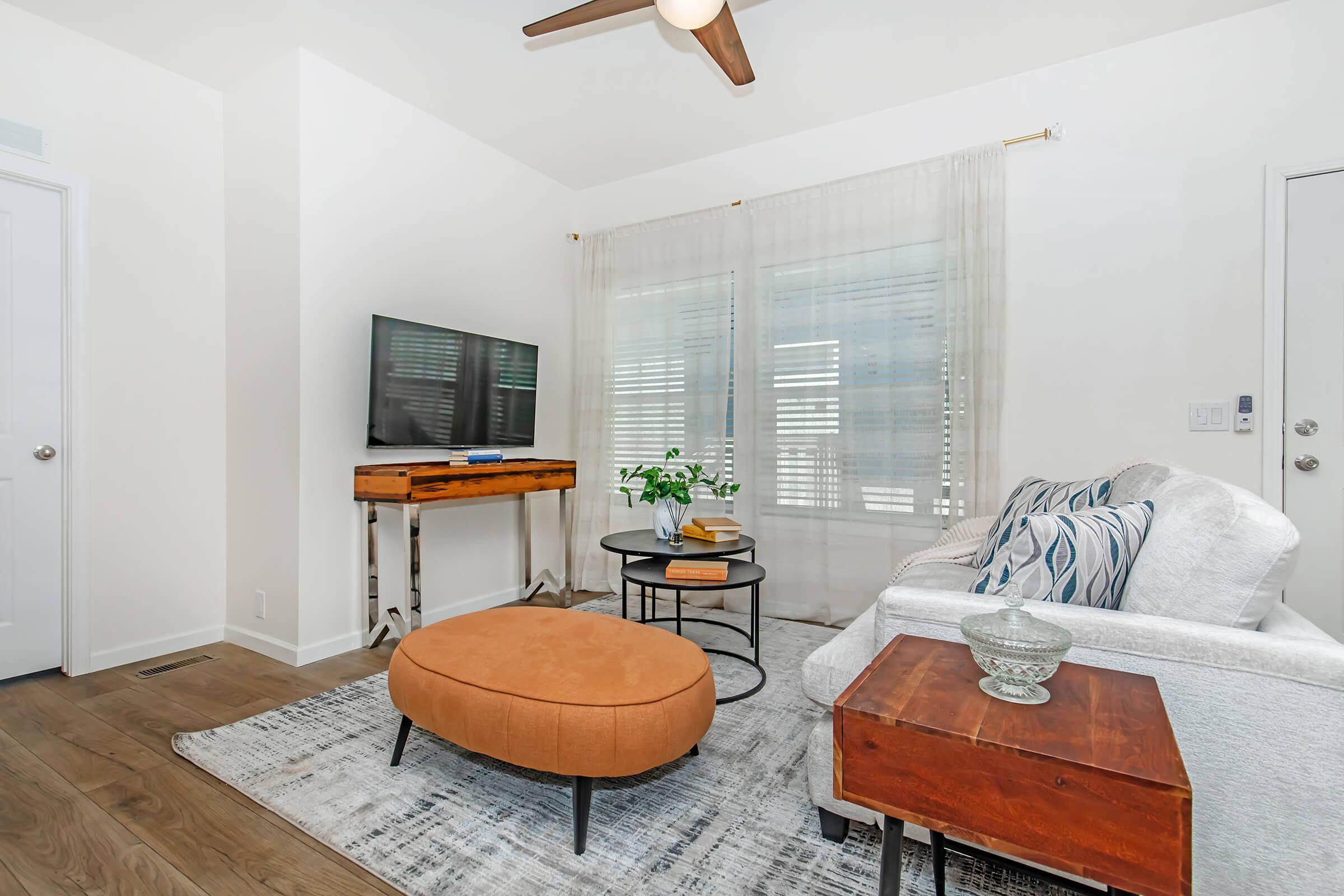A modern living room featuring a plush orange ottoman, a beige sofa with decorative pillows, a wooden coffee table, and a sleek TV mounted on the wall. Light-filtering drapes allow natural light, enhancing the airy atmosphere. Wooden flooring complements the cozy decor, creating a stylish and inviting space.
