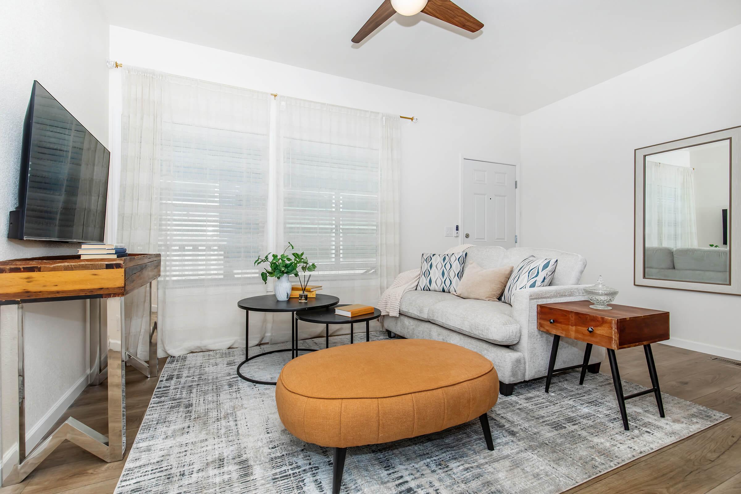 A cozy living room featuring a light gray sofa with decorative pillows, an orange ottoman, and a wooden side table. A modern coffee table sits on a patterned rug. There is a wall-mounted TV, a potted plant, and large windows with sheer curtains, creating a bright and inviting atmosphere.