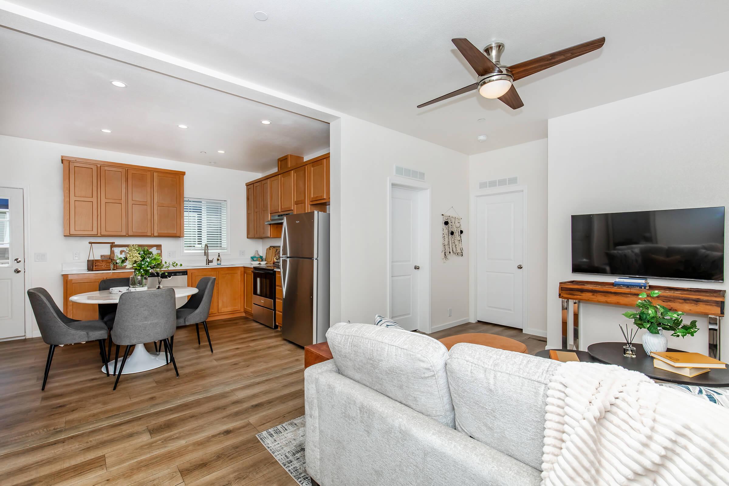 A modern interior of a cozy living space featuring a light gray sofa with a blanket, a wooden coffee table with decor, and a sleek TV on a wooden stand. The open kitchen has wooden cabinets, stainless steel appliances, and a circular dining table with gray chairs. Windows allow natural light to brighten the area.
