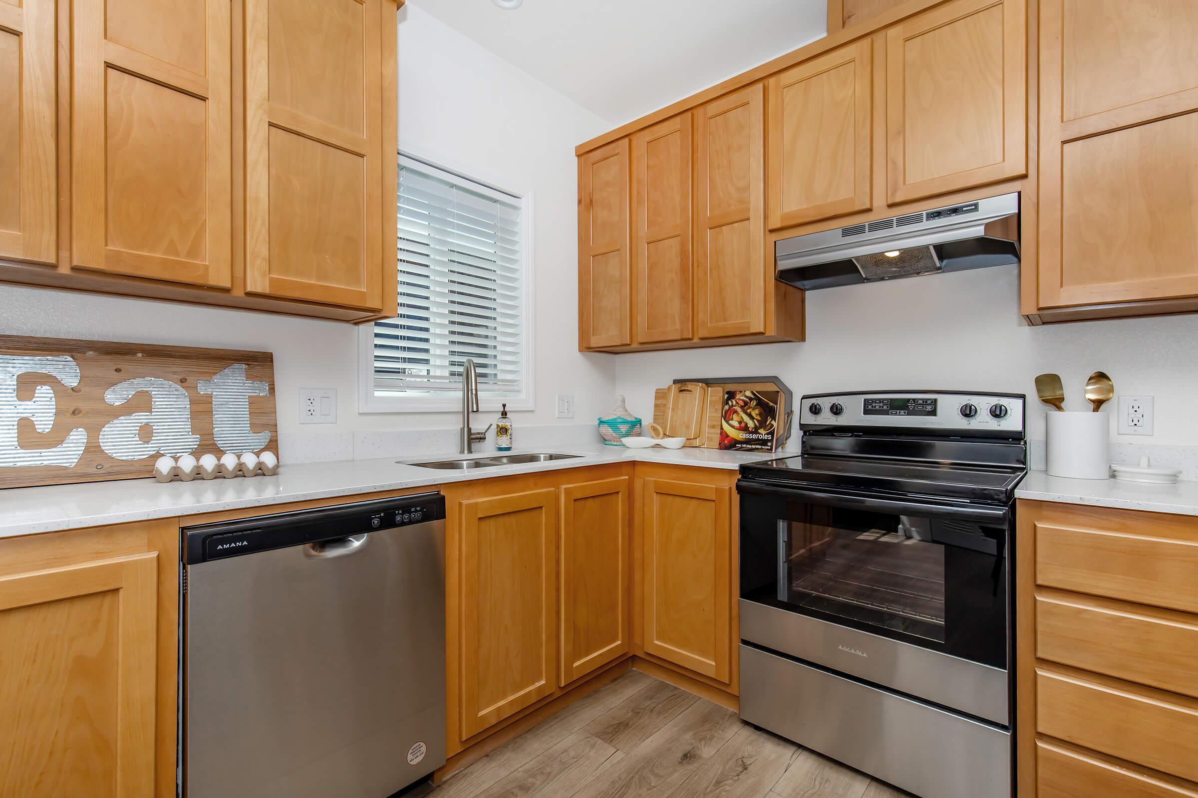 Modern kitchen featuring light wooden cabinetry, a stainless steel dishwasher, an oven, and a sleek stove hood. The countertops are white, and there's a window with white blinds. Decorative accents include a sign that says "Eat" and kitchen tools on display. The flooring is a warm wood tone.