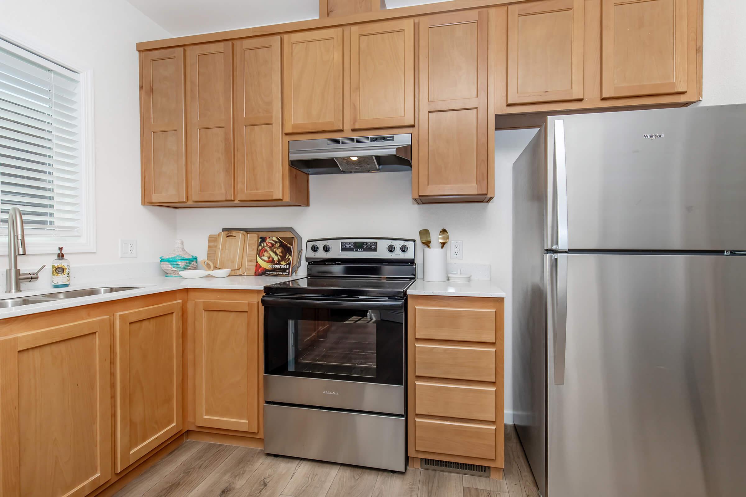 A modern kitchen featuring wooden cabinets, a stainless steel refrigerator, an electric oven with a glass cooktop, a sink, and various kitchen items on the counter. The space has light-colored flooring and a window with blinds, allowing natural light to enter.
