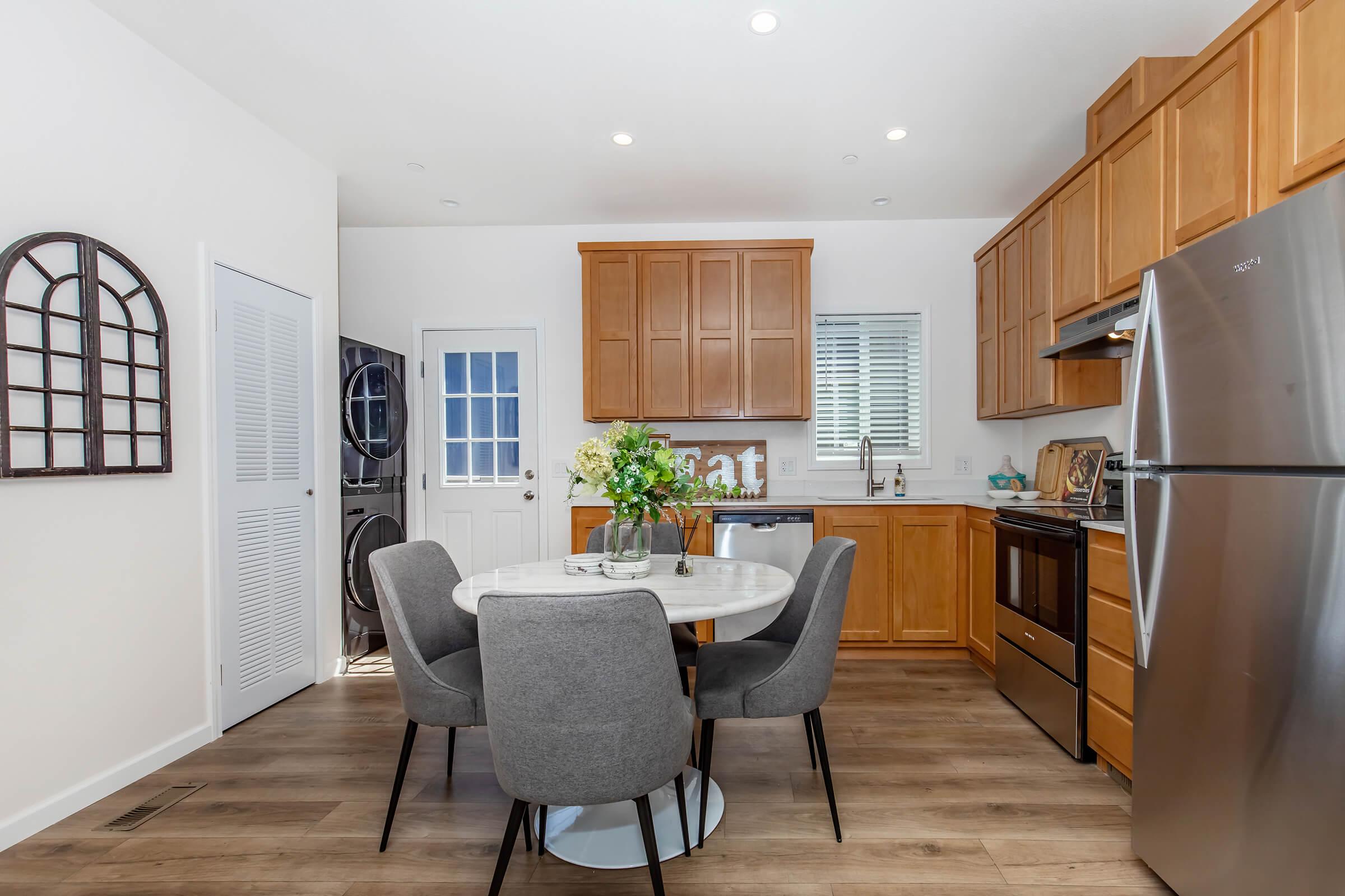 A modern kitchen featuring wooden cabinetry, stainless steel appliances, and a round dining table with four gray chairs. The space is well-lit with natural light, and there is a door leading outside. A vase with flowers is placed on the table, adding a touch of decor.