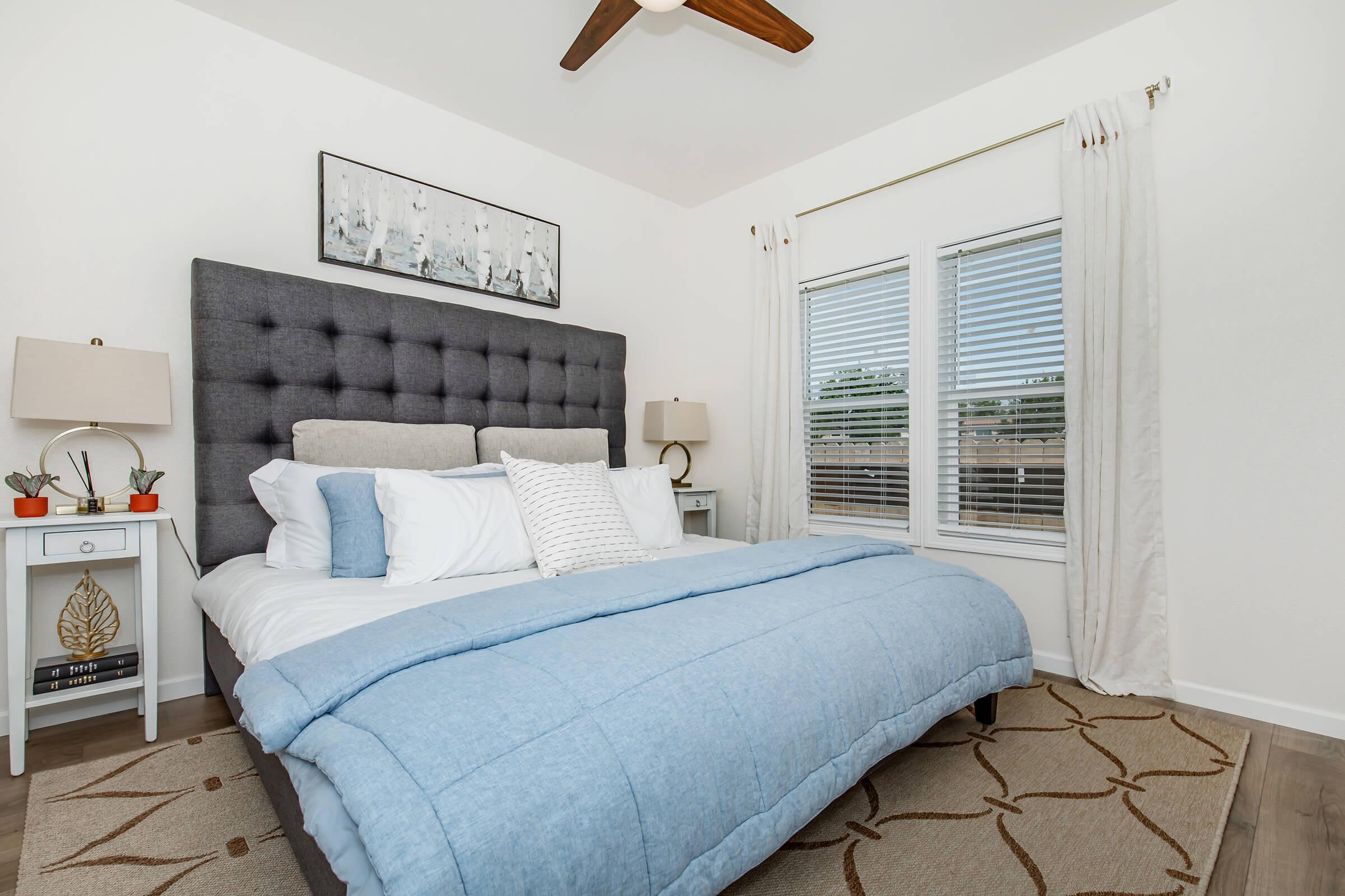 A bright and modern bedroom featuring a large tufted gray headboard, a neatly made bed with white and light blue bedding, and two bedside tables with lamps. Natural light streams through white curtains covering the window, and a neutral-toned area rug adds warmth to the wooden floor.