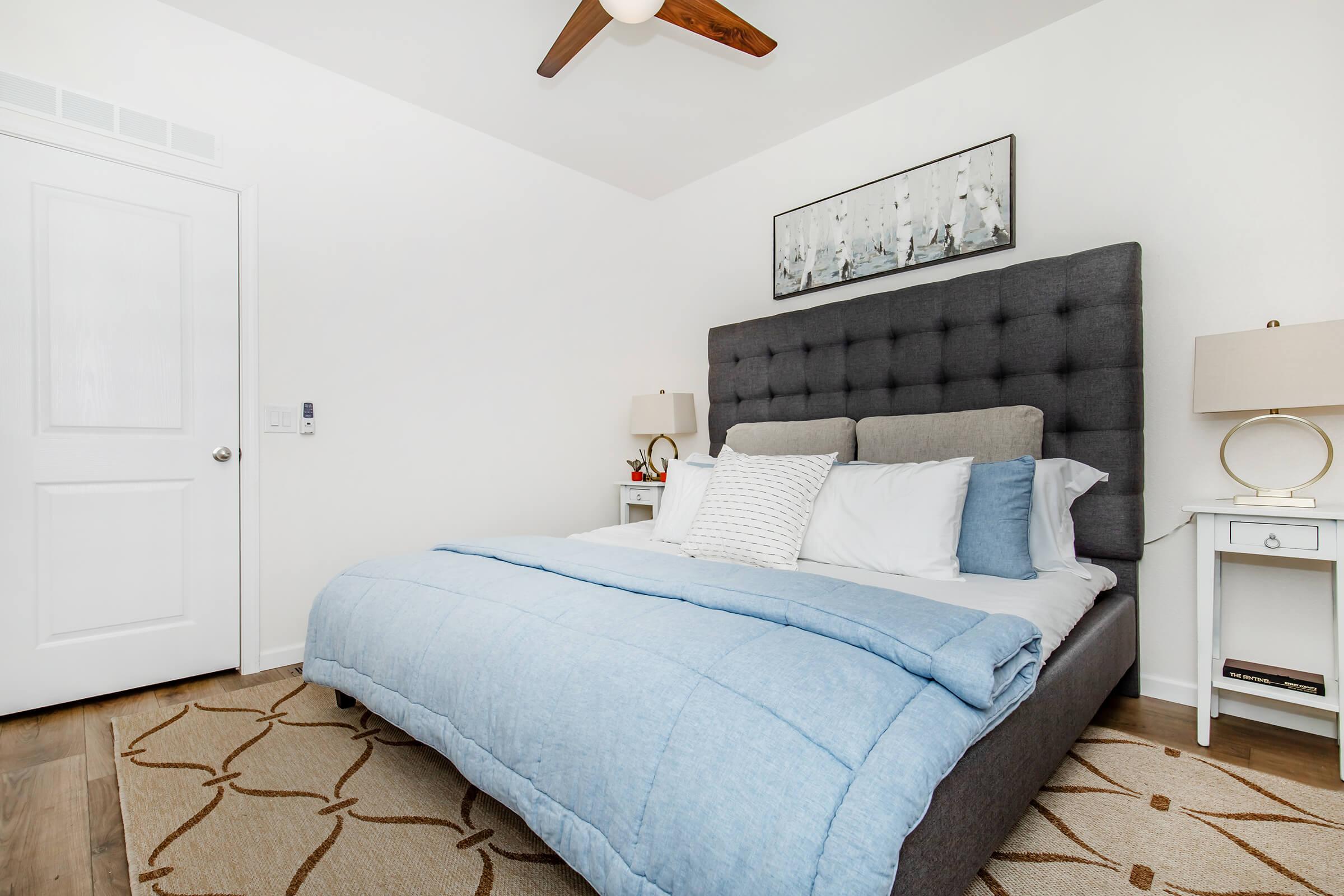 A cozy bedroom featuring a large gray upholstered bed with blue bedding, two bedside tables with lamps, a ceiling fan, and a decorative wall art piece. The walls are painted white, and the floor is covered with a light-colored area rug displaying a geometric pattern.