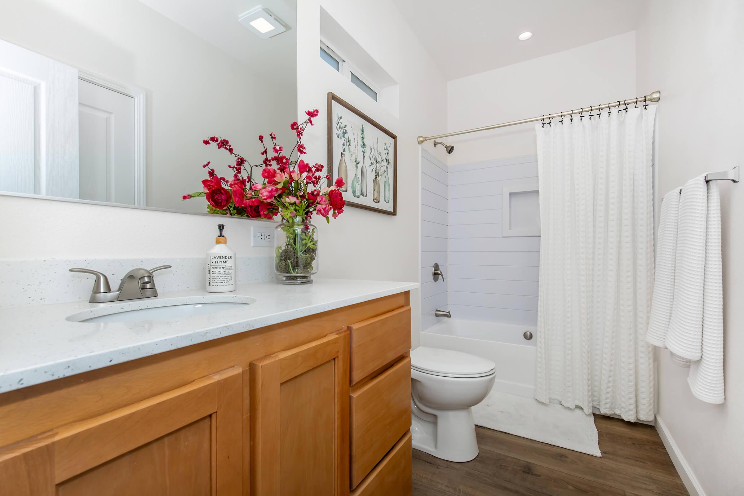 Bright and modern bathroom featuring a vanity with a sink and wooden cabinetry. A shower with a white curtain and a small window is visible. A vase with vibrant red flowers adds a pop of color, while plush towels hang by the toilet. The space has a clean, fresh aesthetic with light walls and flooring.