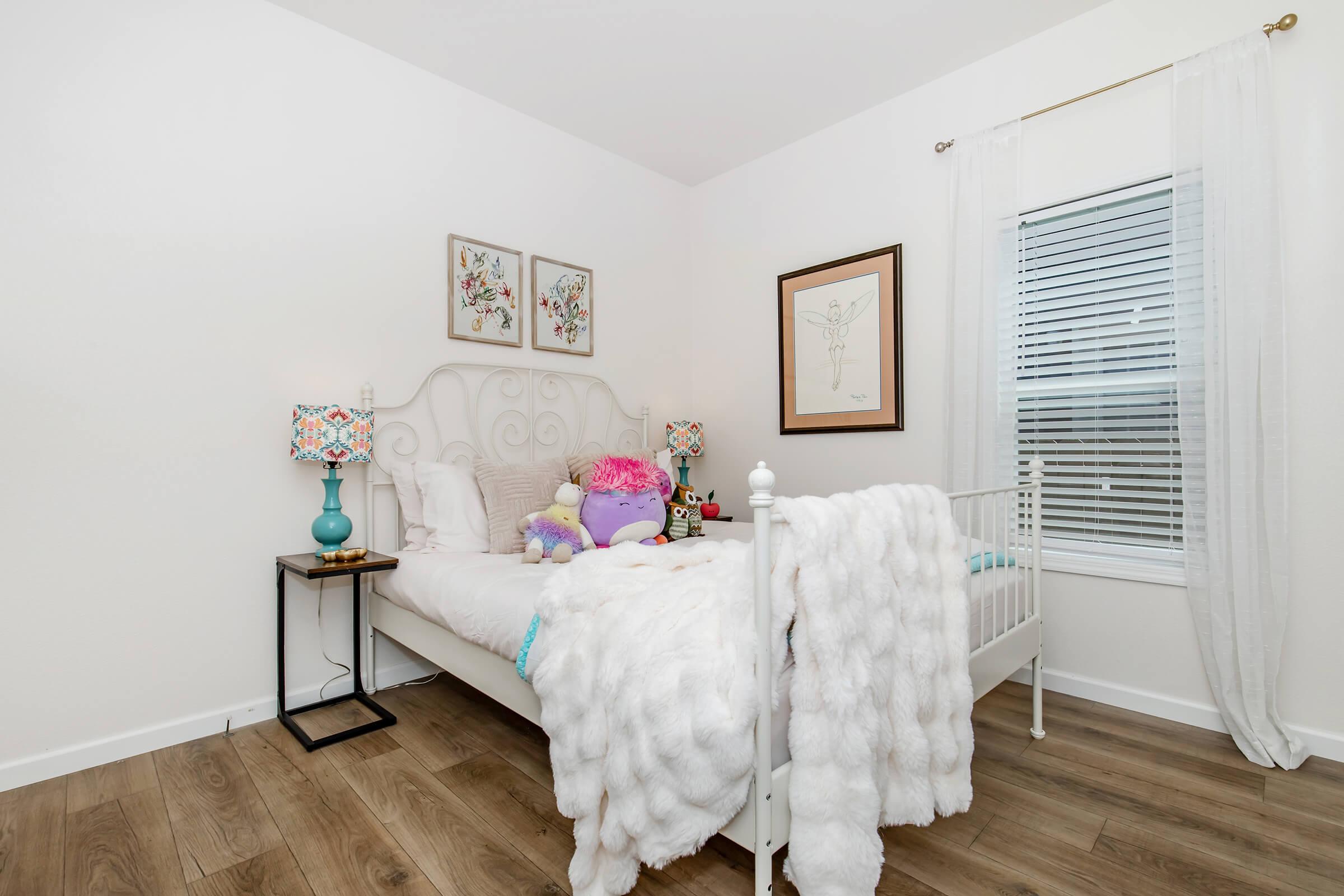 A cozy bedroom featuring a white metal bed with a fluffy blanket, colorful decorative pillows, and plush toys. There are two bedside lamps with floral designs and framed artwork on the wall. The room has light wood flooring and a window with sheer curtains allowing natural light to enter.