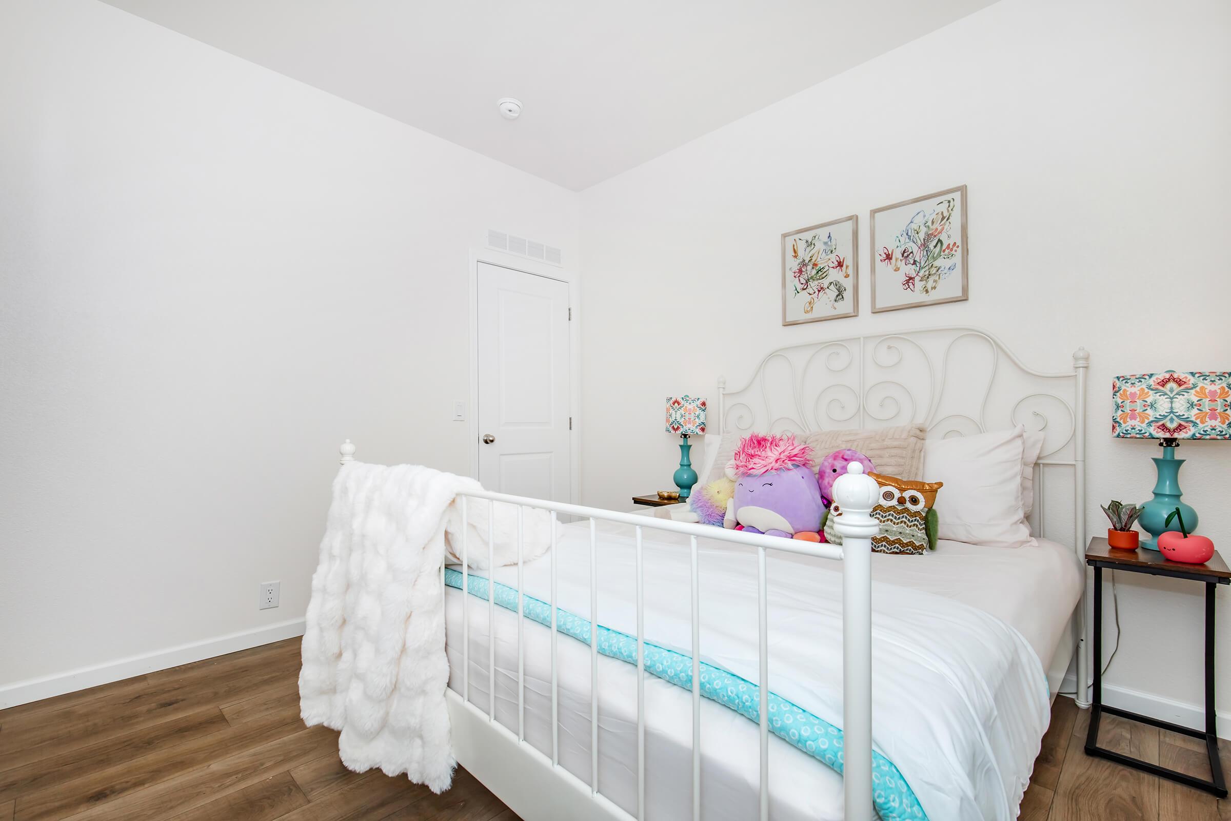 A cozy bedroom featuring a white bed with colorful pillows, a fluffy throw blanket draped over the side, two bedside lamps with vibrant shades, and light wood flooring. The walls are painted white, and there are framed floral artworks above the bed, creating a cheerful and inviting atmosphere.