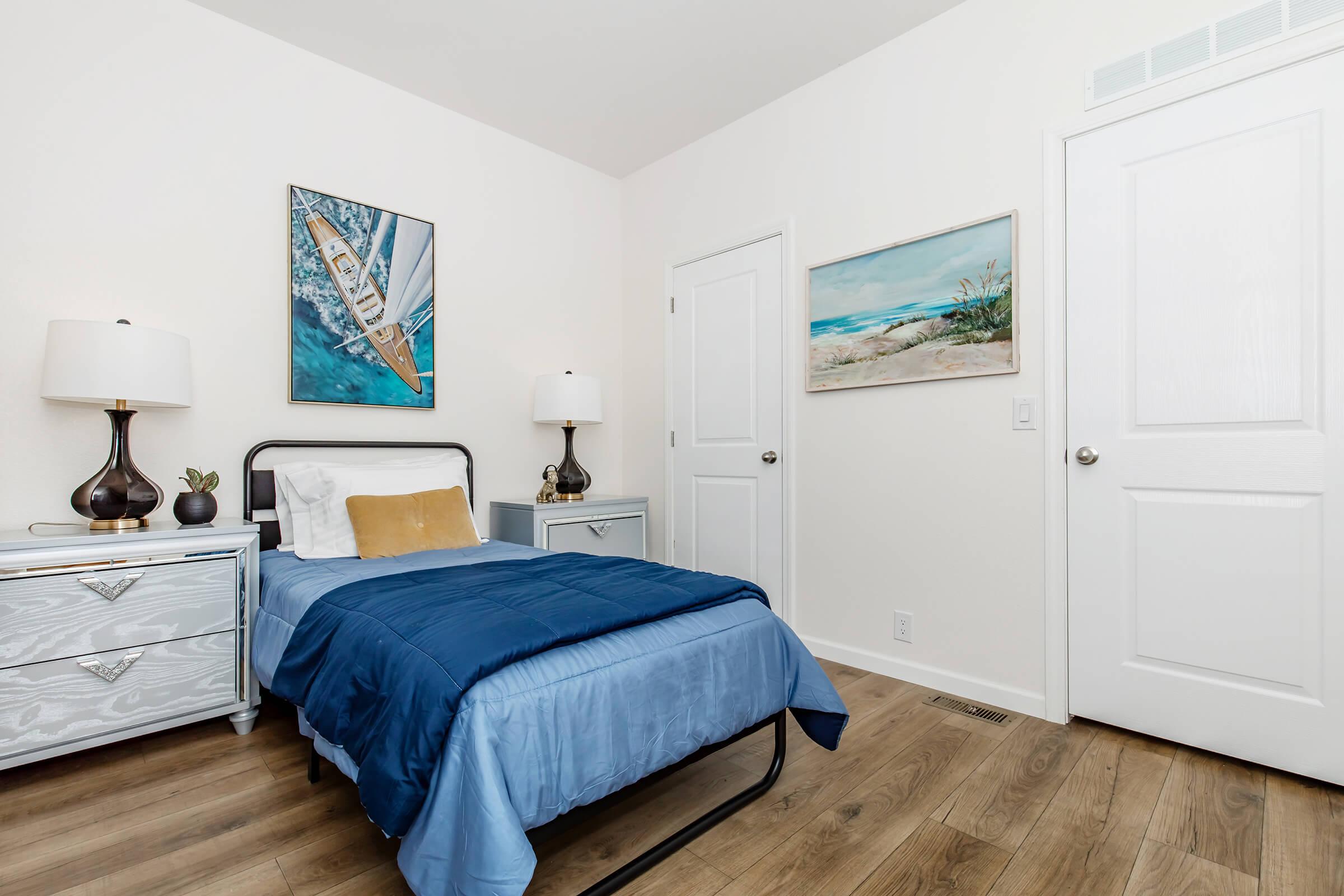 A modern bedroom featuring a single bed with a blue comforter, two bedside tables with lamps, and artwork on the walls depicting a sailing scene and a beach. The room has light-colored walls and wooden flooring, creating a bright and inviting atmosphere. A closed door is visible on the right.