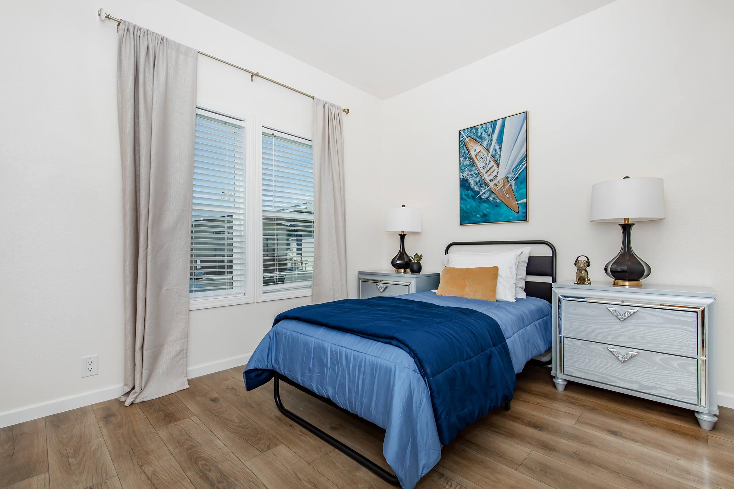 A cozy bedroom featuring a single bed with a blue comforter, a decorative pillow, and a bedside table with a lamp. There is an artwork of a sailboat on the wall, a window with blinds allowing natural light, and light-colored walls and wooden flooring, creating a tranquil atmosphere.