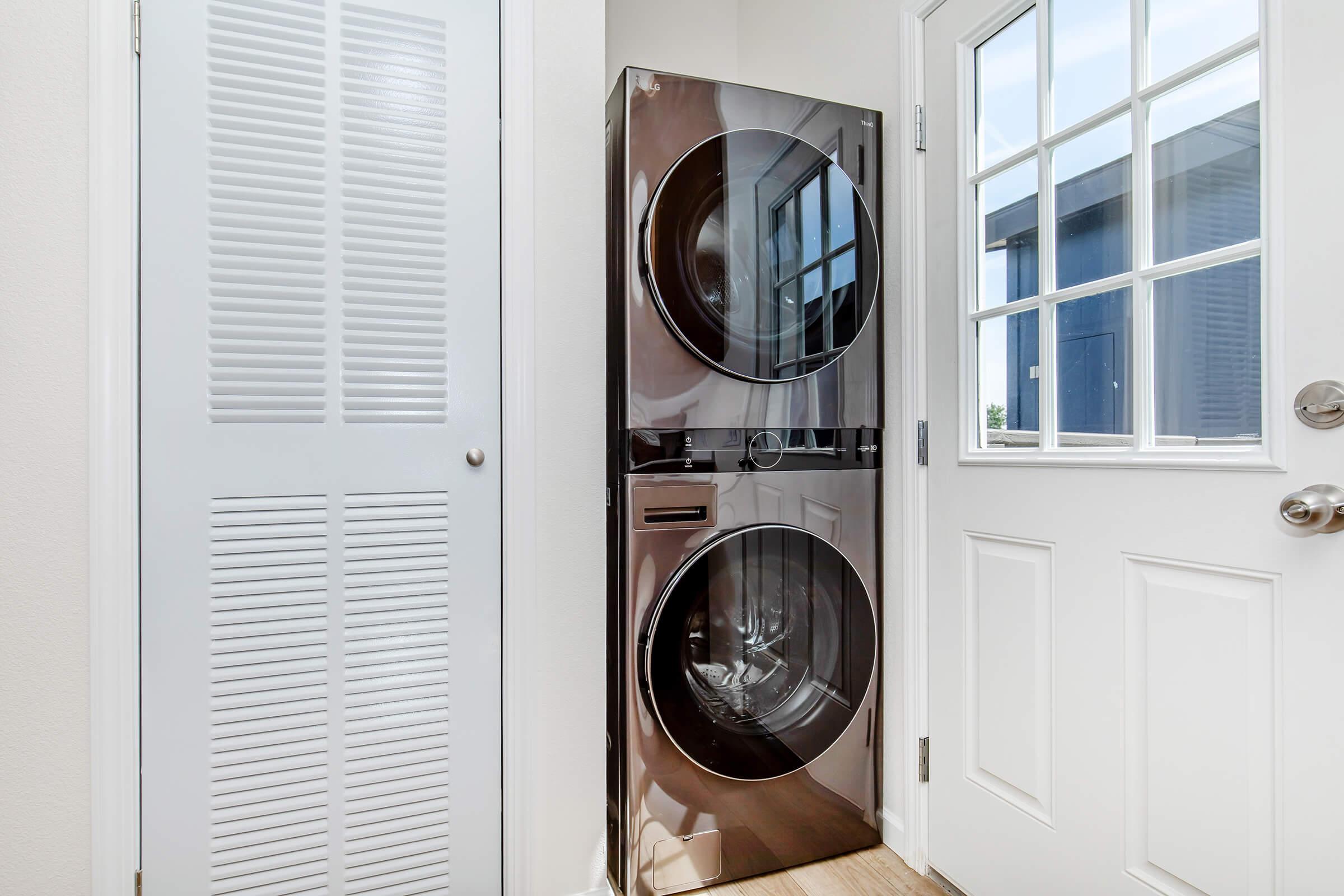 A modern laundry area featuring a stacked black washer and dryer unit next to a white door with glass panels. The space has light-colored walls and a closet door with vertical slats, providing a clean and organized look. Natural light filters in through the door, enhancing the brightness of the area.