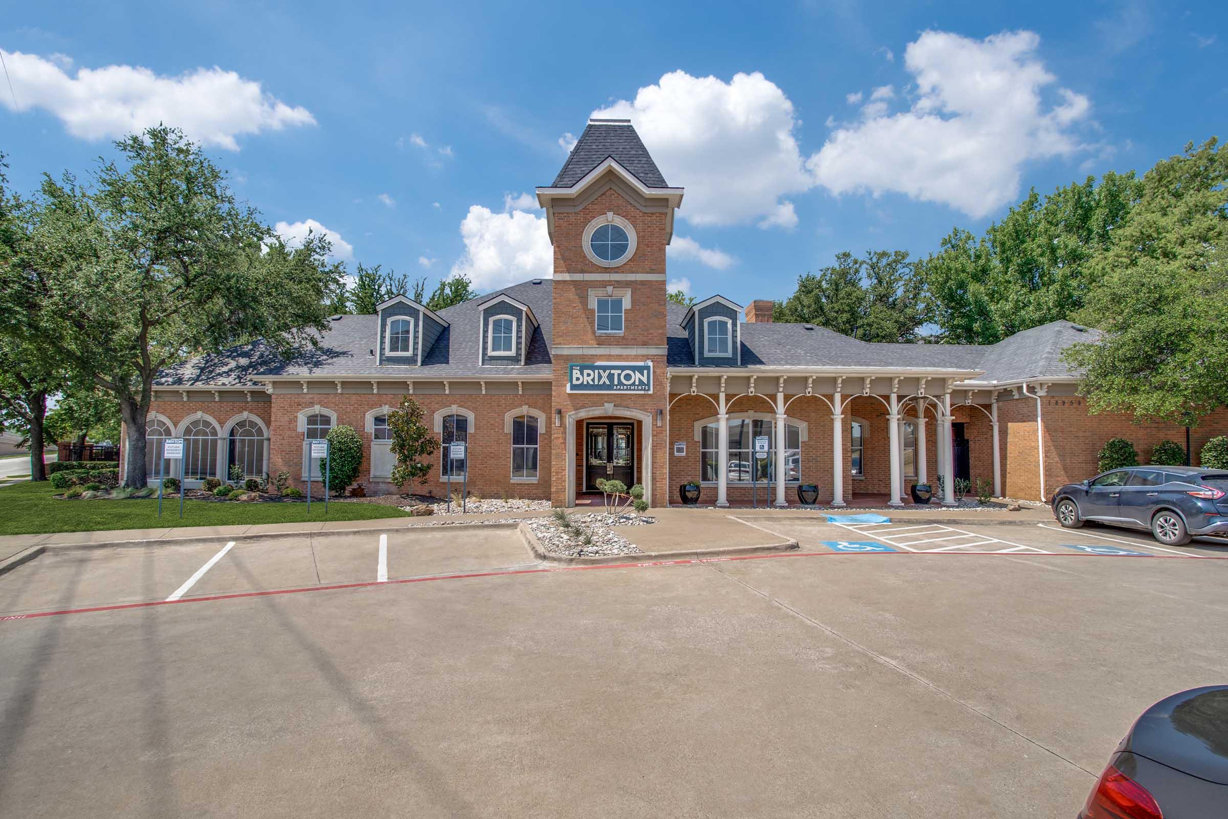 A two-story brick building with a clock tower and a sign that reads "BRIXTON." The architecture features arched windows and a covered porch. Surrounding landscaping includes trees and shrubs, with parked cars in the foreground and a clear blue sky with fluffy clouds above.