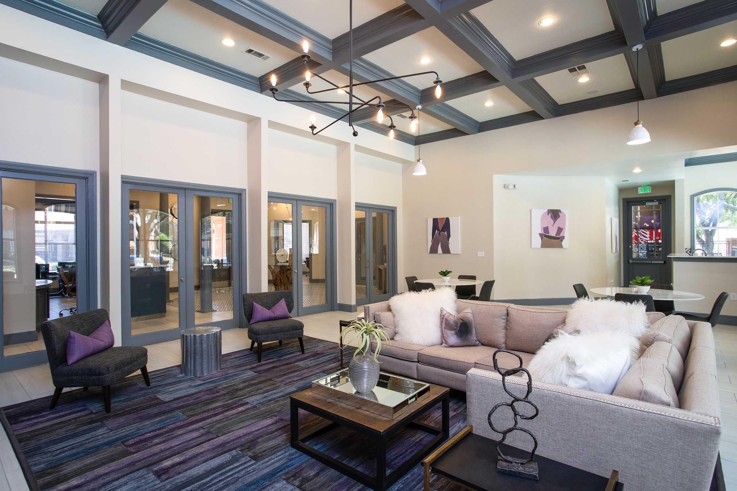 A modern, stylish living area featuring a light-colored sofa with decorative pillows, a coffee table, and a plush rug. The ceiling has dark wooden beams, and there are two elegant chairs nearby. Walls are painted in soft tones, complemented by framed artworks and large windows letting in natural light.