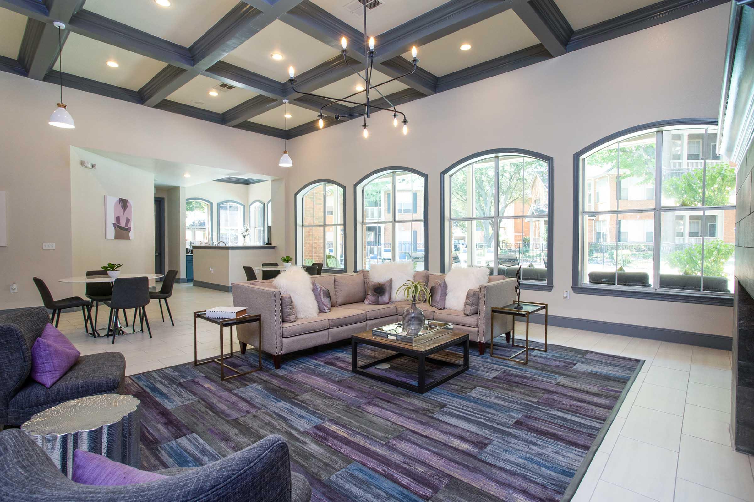 A cozy living room featuring a modern sofa, accent chairs, and a coffee table on a colorful area rug. Large arched windows allow natural light, showcasing greenery outside. The space has stylish lighting fixtures and a contemporary decor, creating an inviting atmosphere.
