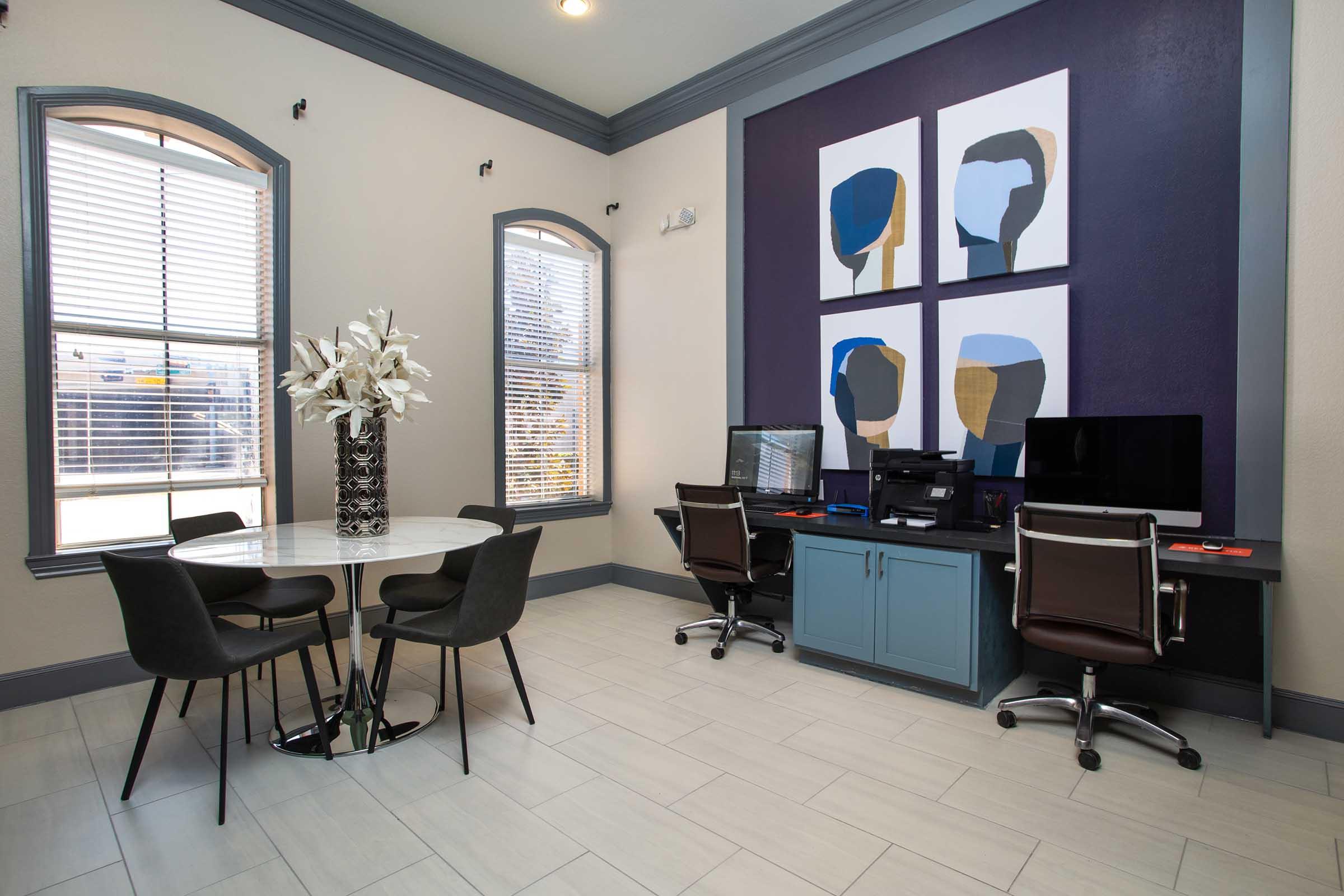 Stylish office space featuring a round dining table with four black chairs, a large vase of white flowers, and two computer desks with black chairs. The walls are painted in light colors, accented with modern artwork depicting abstract figures. Natural light comes through large windows with blinds.