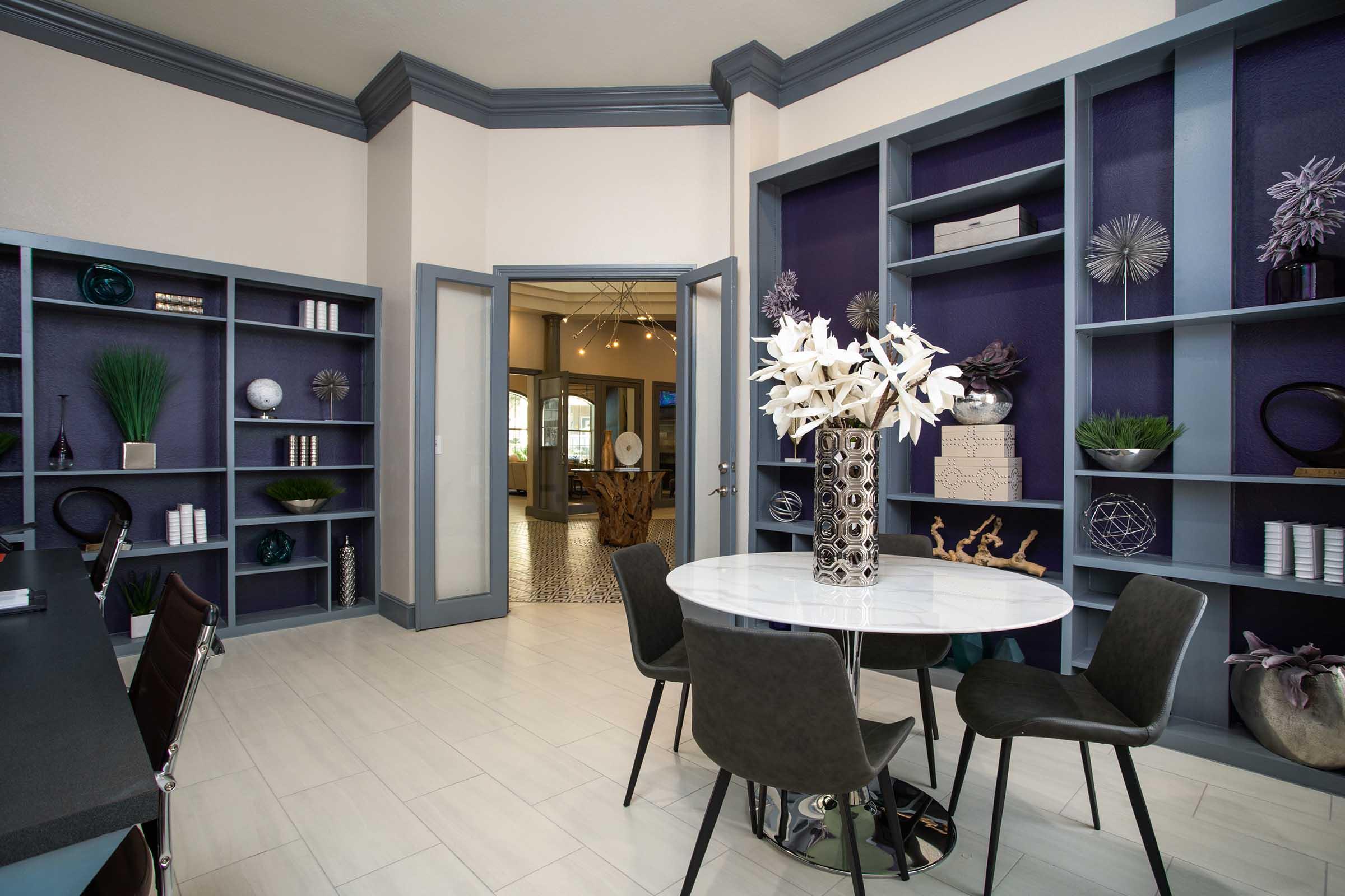 A modern interior space featuring a round dining table surrounded by black chairs. The room has blue-gray walls with open shelving displaying decorative items and plants. One wall is painted deep purple, adding contrast to the light-colored flooring. A doorway leads to another room in the background.