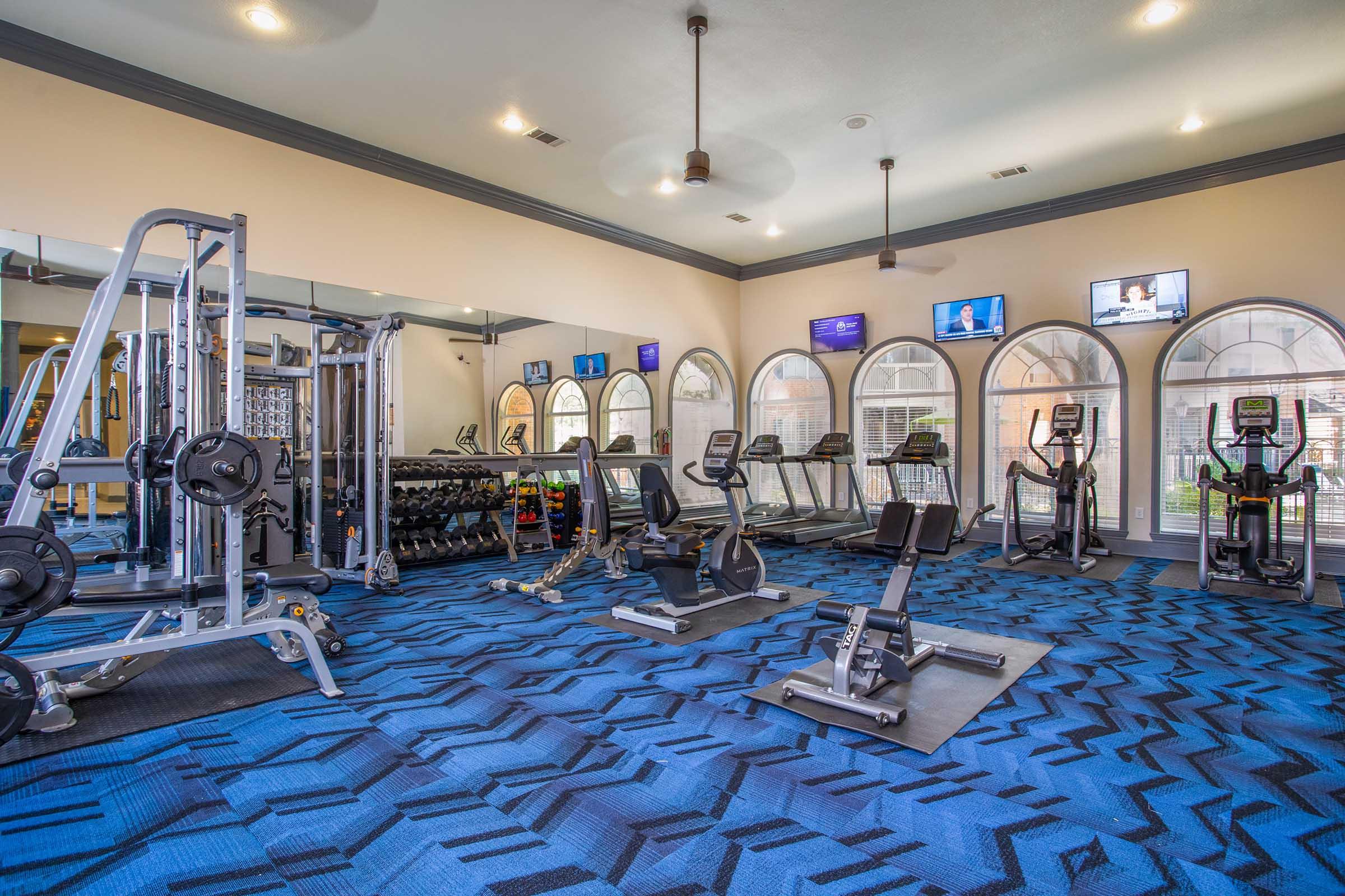 A modern gym interior featuring various exercise equipment, including treadmills, stationary bikes, weight machines, and free weights. The space has large arched windows allowing natural light and is decorated with a blue patterned carpet. Several TVs are mounted on the walls, displaying news channels.