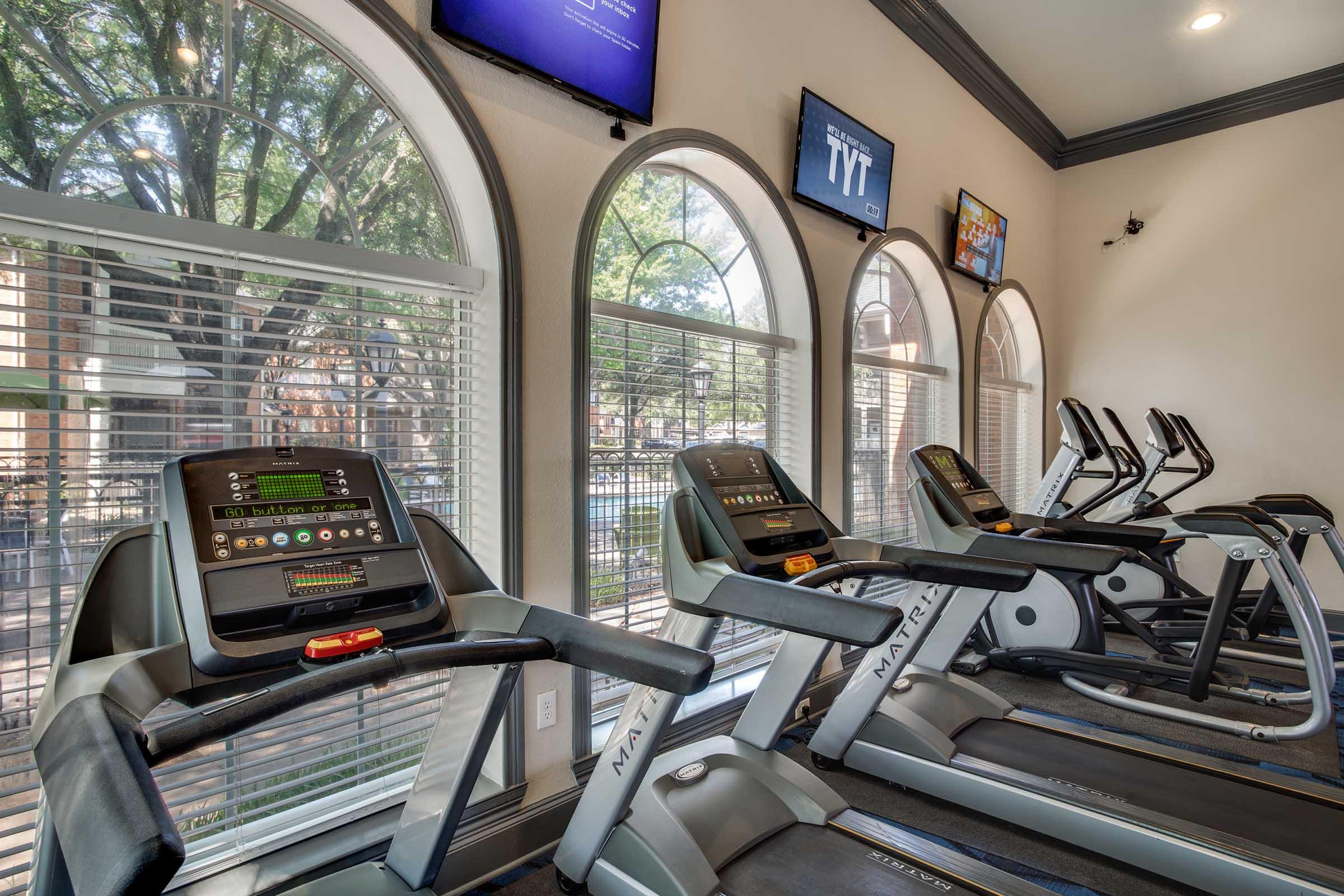 A bright and spacious gym with several treadmills and elliptical machines lined up against large arched windows. Natural light streams in, illuminating the equipment, while screens display workout programs and information. The environment is clean and modern, creating an inviting space for exercise.