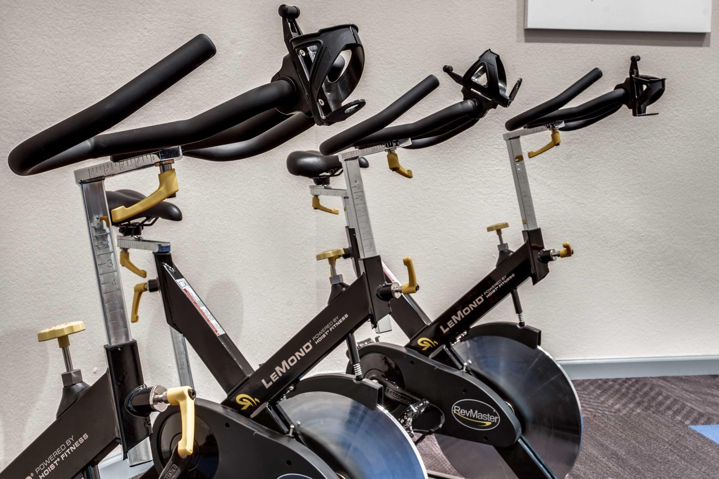 Row of stationary spin bikes in a gym setting, featuring adjustable handlebars and sturdy frames. The bikes are positioned on a carpeted floor, suggesting a fitness environment. Bright lighting enhances the space.