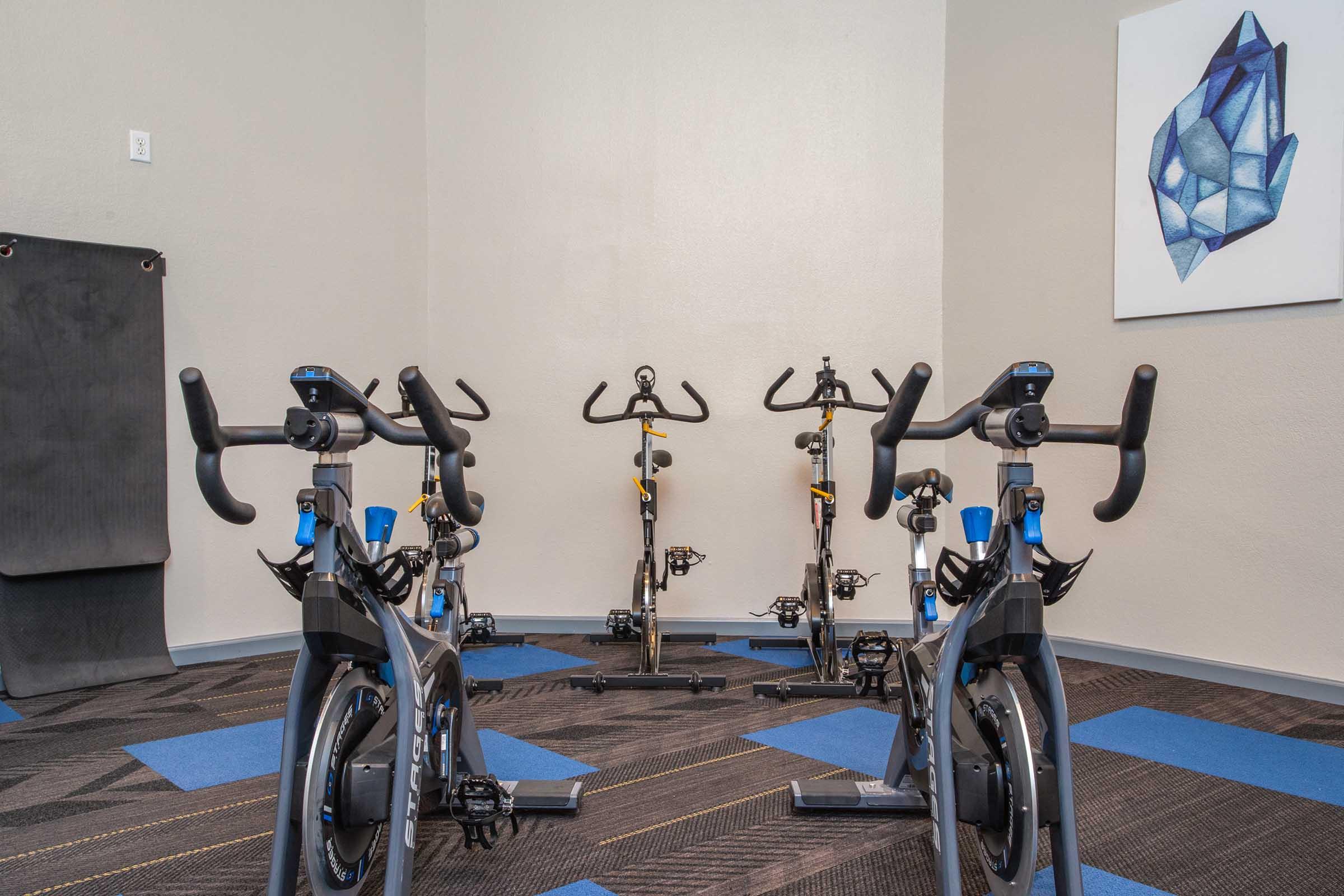 A fitness studio featuring multiple stationary bikes arranged in a corner. The room has gray walls, blue and black flooring, and a geometric artwork on the wall. The setup is designed for indoor cycling workouts.