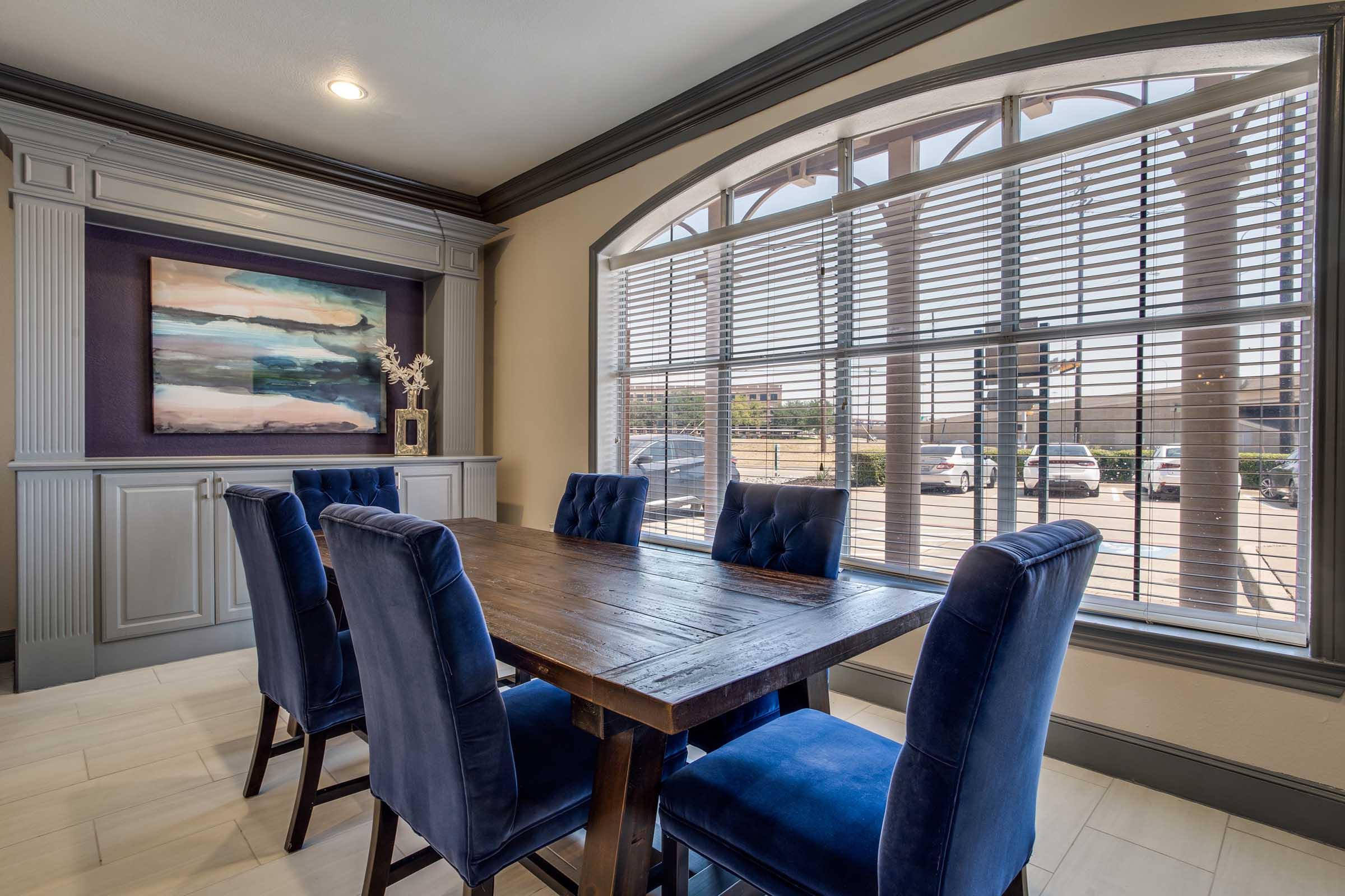 A modern dining area featuring a rectangular wooden table surrounded by blue upholstered chairs. A large window with blinds allows natural light to fill the space. A colorful abstract painting hangs on the wall above a built-in cabinet, adding a touch of decor to the room.