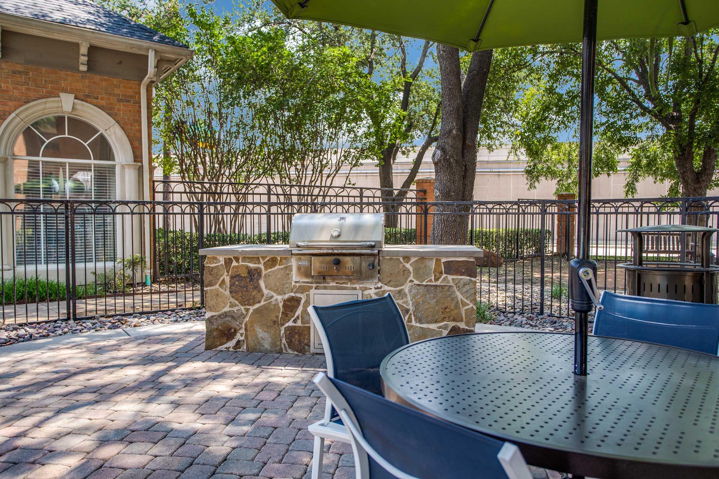 Outdoor patio area featuring a stone barbecue grill, surrounded by trees and a decorative fence. A round, metal table with chairs is positioned nearby, under a green umbrella. The setting suggests a relaxed atmosphere for outdoor gatherings or dining.