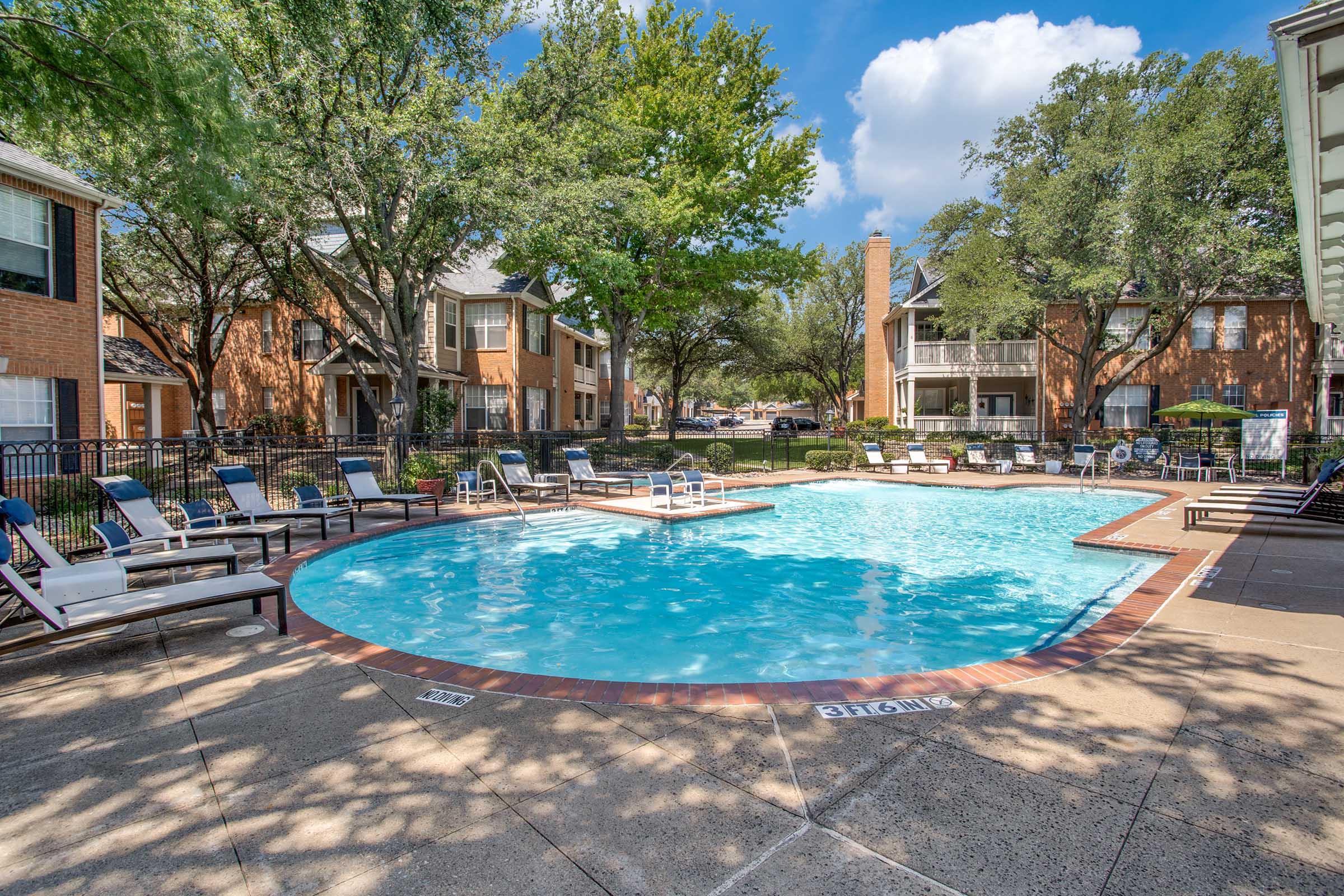 A clear blue swimming pool surrounded by lounge chairs in a residential area. Lush green trees provide shade, and nearby buildings feature brick exteriors. The sky is partly cloudy, adding to the serene atmosphere of the outdoor space.