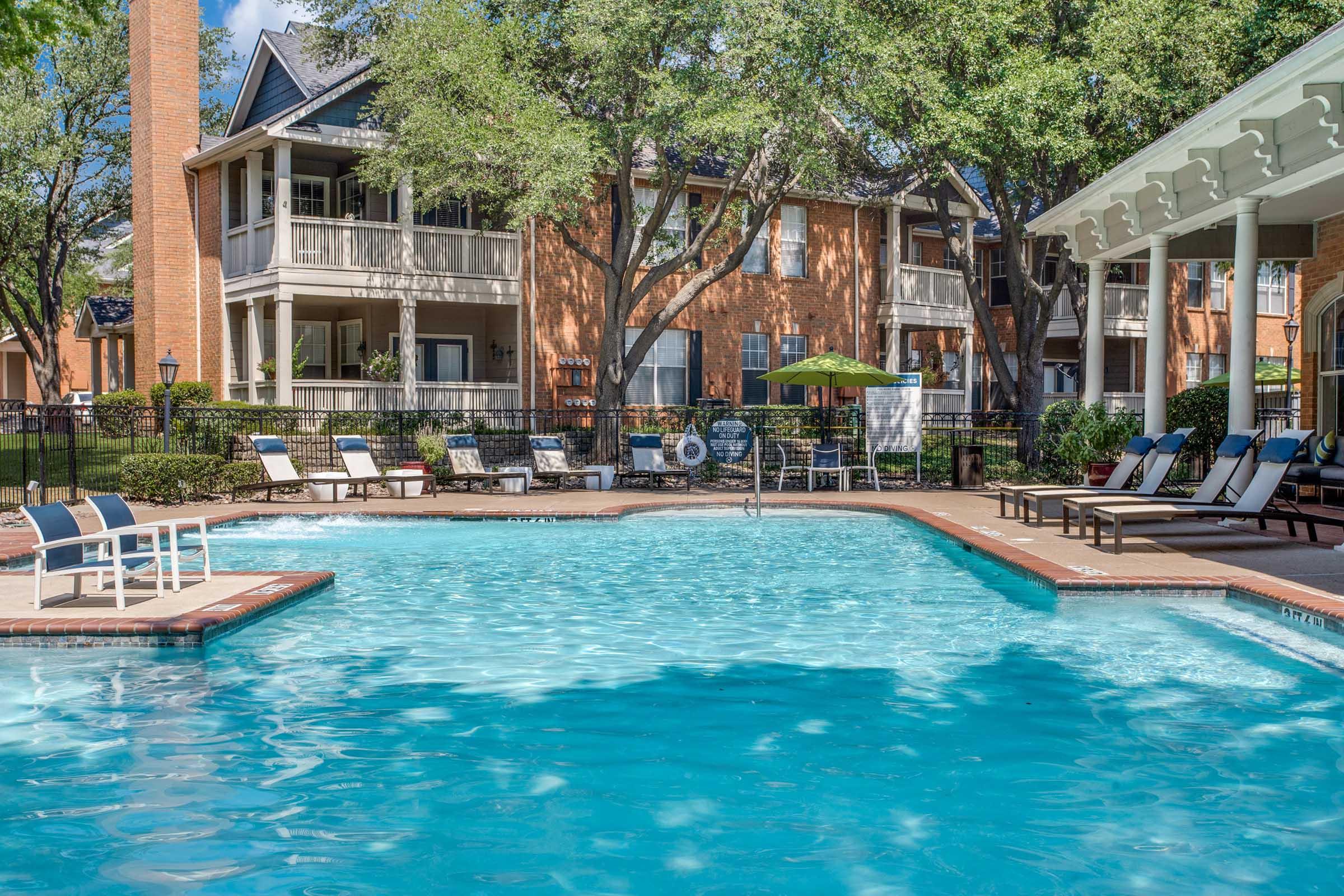 A sunny outdoor pool area surrounded by lounge chairs and umbrellas, with trees providing shade. In the background, brick buildings with balconies create a cozy residential atmosphere. The water appears clear and inviting, ideal for relaxing or socializing.