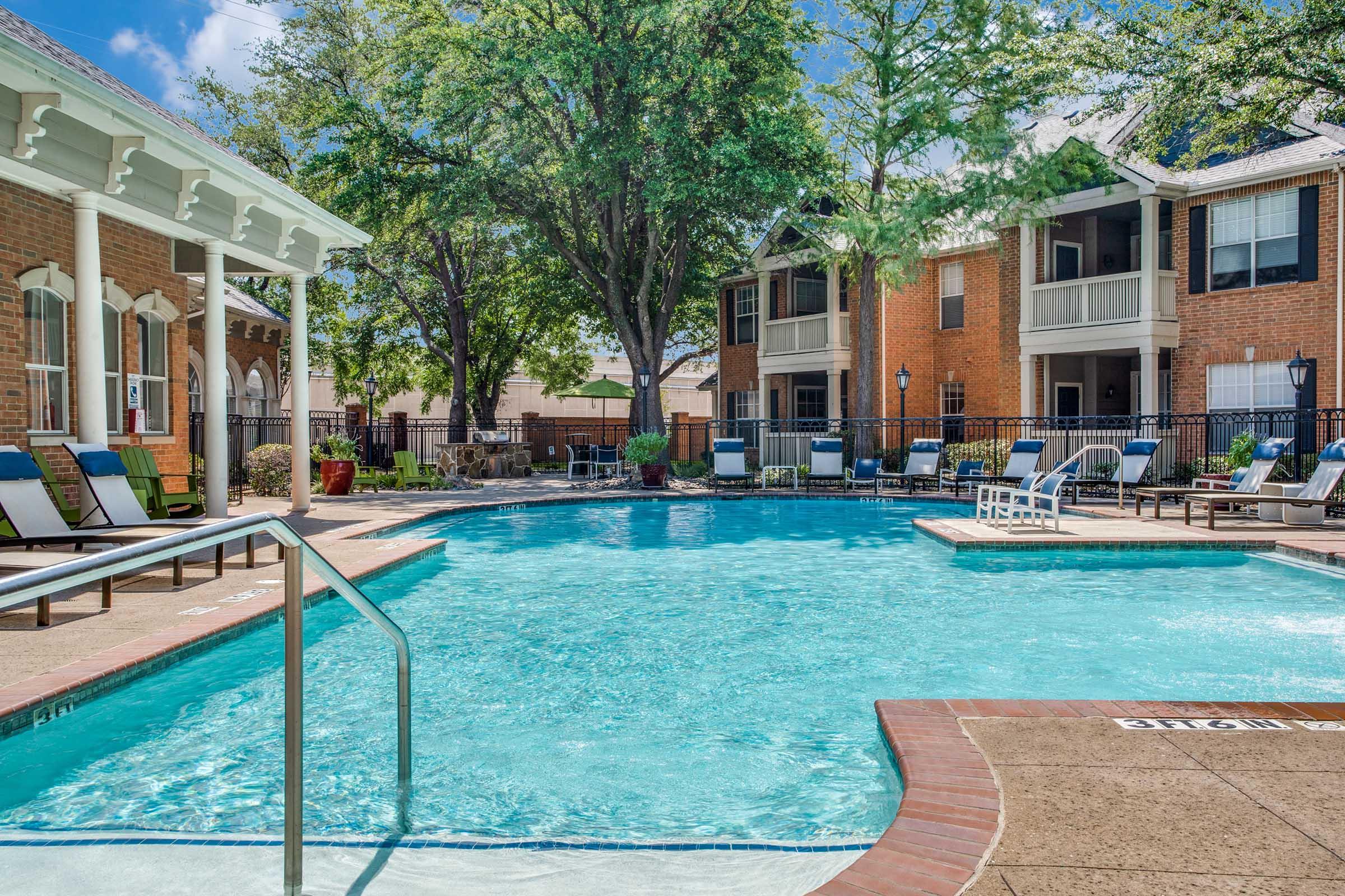 A bright outdoor swimming pool surrounded by lounge chairs and leafy trees. Apartment buildings with balconies can be seen in the background. The water is clear and inviting under a sunny sky, creating a relaxed and inviting atmosphere.