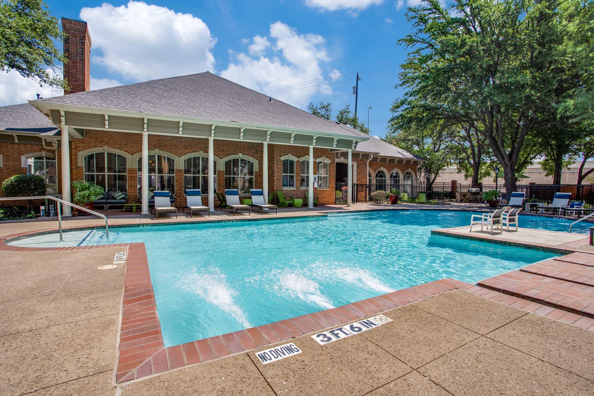 A clear blue swimming pool surrounded by a patio. Lounge chairs line the edges, and trees provide shade in the background. The pool features a deep end, labeled at 3 feet, 6 inches. A brick wall and a building with large windows are visible near the pool area under a bright blue sky with fluffy clouds.