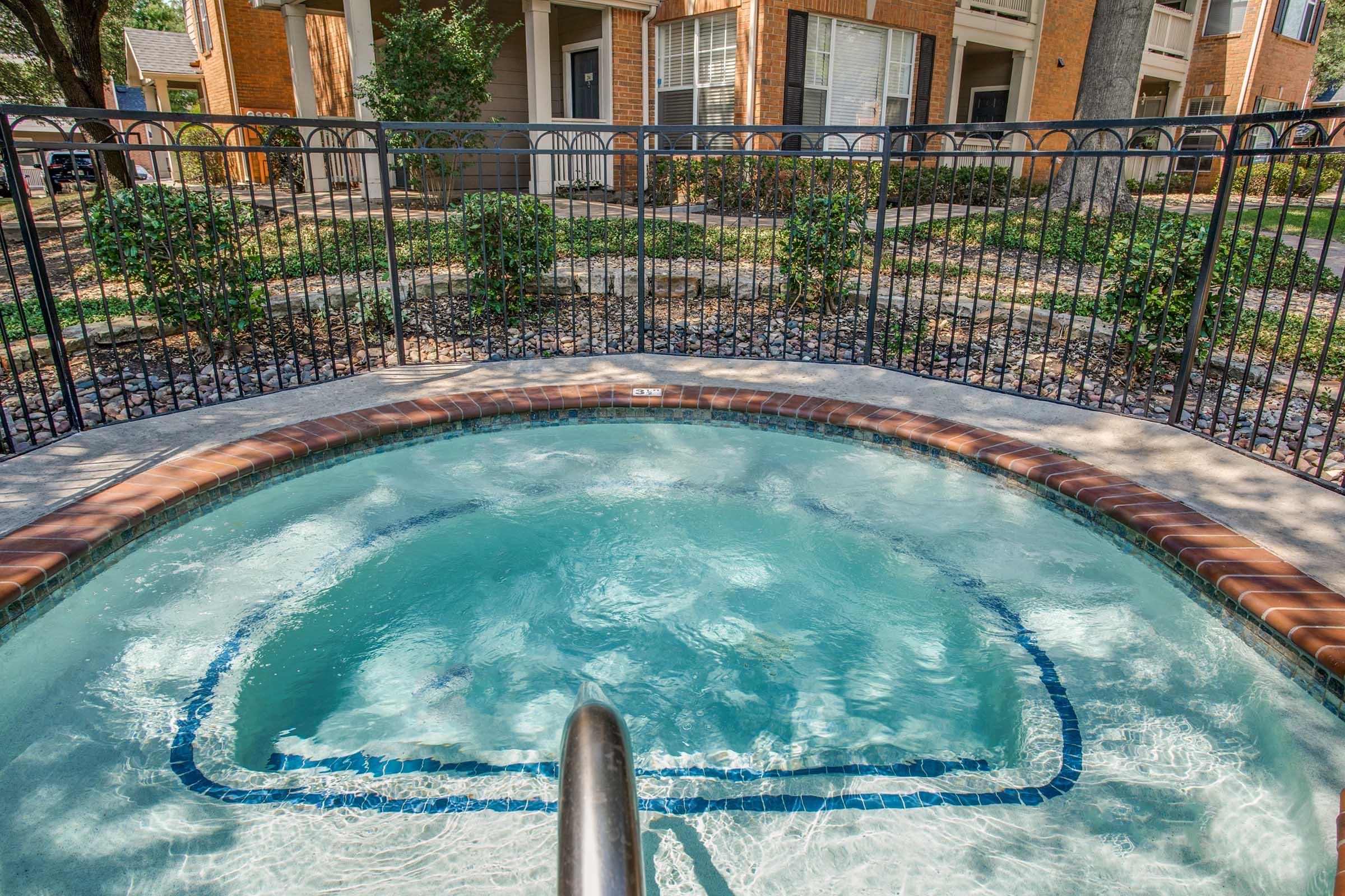 A view of a hot tub with bubbling water, surrounded by a well-maintained patio, partial fencing, and neatly arranged shrubs. In the background, there are residential buildings with balconies and windows, indicating a multi-unit living area. Sunlight is casting gentle reflections on the water.