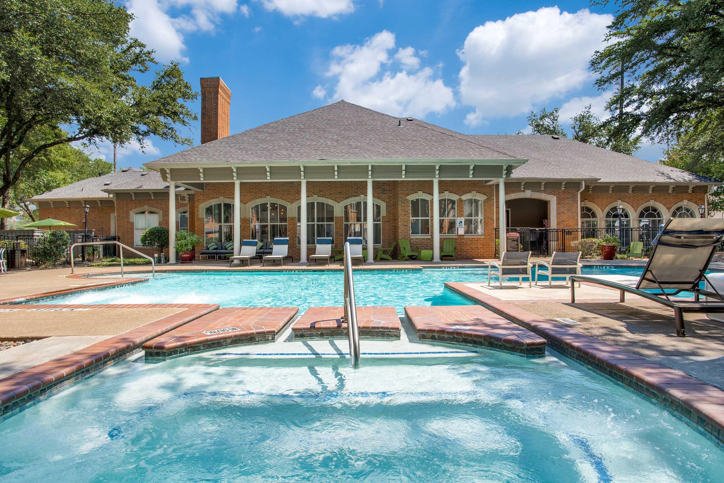 A well-maintained outdoor pool area featuring a large swimming pool and a hot tub. The pool is surrounded by comfortable lounge chairs and shaded areas, with a beautiful brick building in the background. Lush trees and blue skies enhance the relaxing atmosphere.