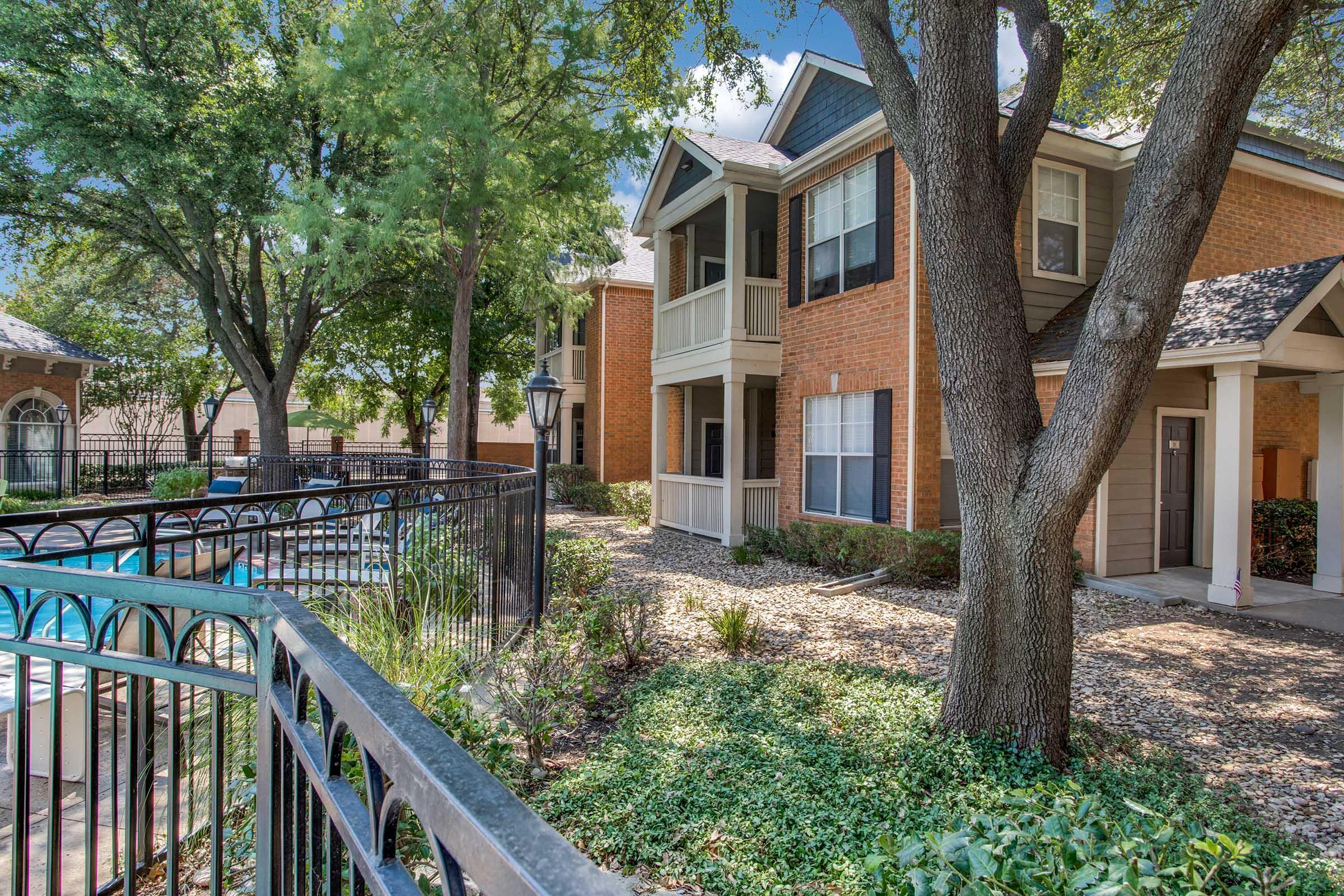 A well-maintained apartment complex featuring brick buildings surrounded by lush greenery and trees. A wrought iron fence encloses a swimming pool area, enhancing the inviting atmosphere. Bright blue skies provide a picturesque backdrop.