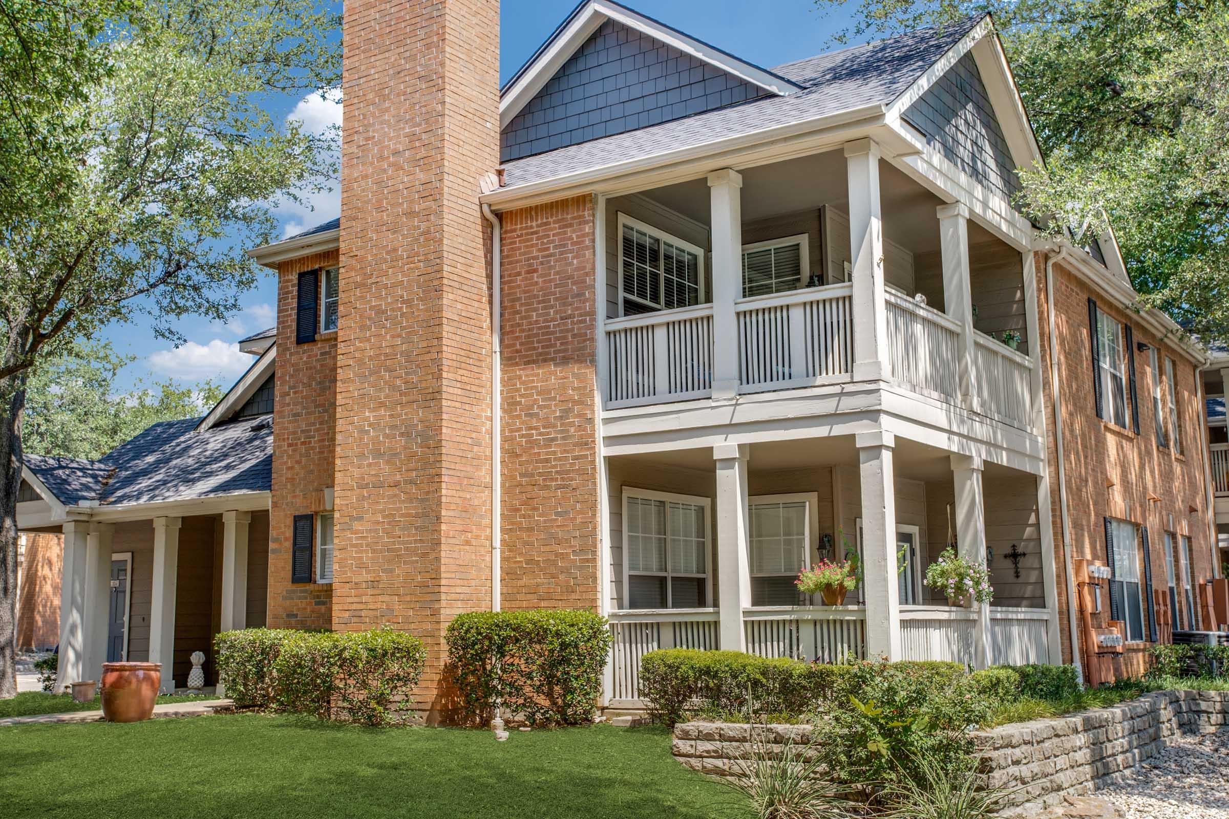 A two-story brick apartment building with a modern design, featuring porches with white railings, lush green landscaping, and a well-maintained garden. The exterior includes a tall chimney and large windows, with sunny weather and blue sky enhancing the inviting atmosphere.