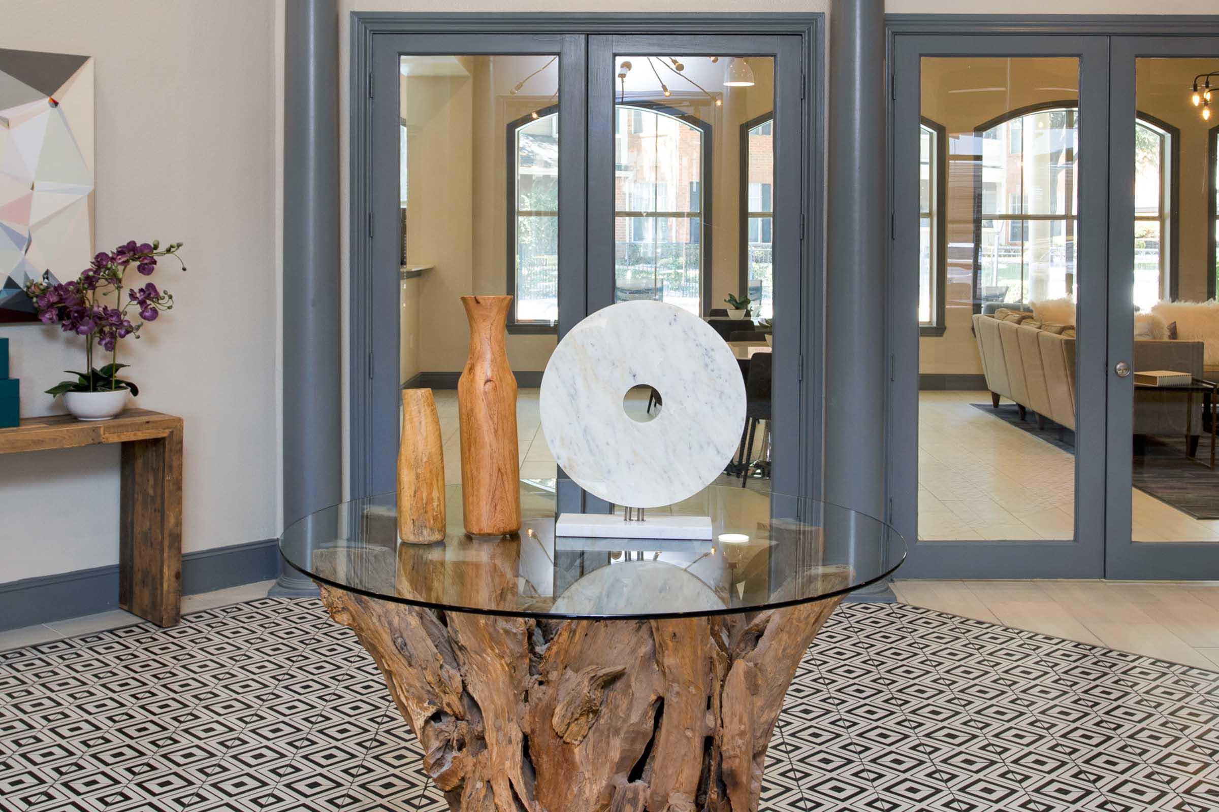 A modern lobby featuring a glass-top table with a wooden base, showcasing three decorative vases in natural wood and white marble. The background includes large arched windows, allowing natural light to illuminate the space, and a patterned floor adds an elegant touch.