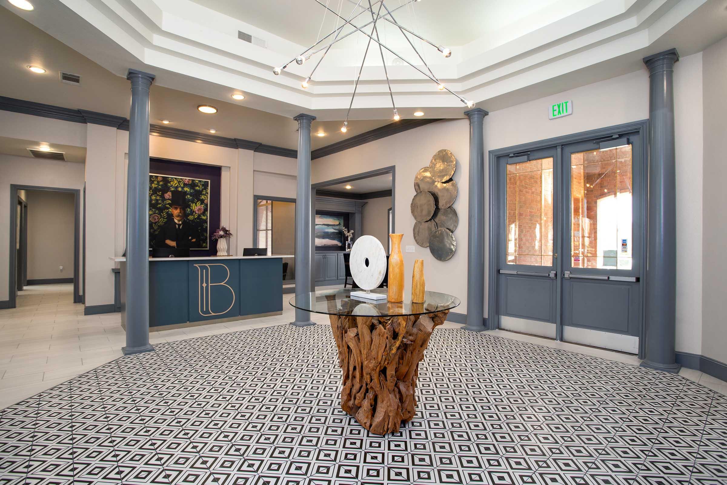 Interior view of a modern lobby featuring a unique wooden table with a circular white sculpture on top. The floor is covered with a black-and-white geometric patterned tile. In the background, a reception desk is visible, along with decorative elements and large windows.