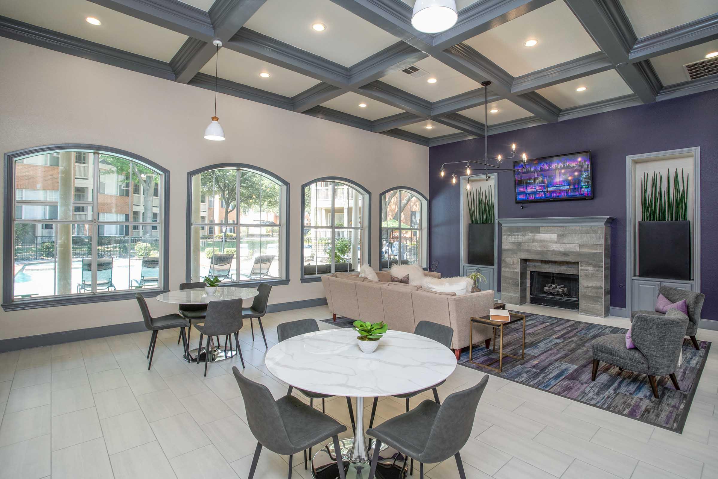 A modern living area featuring large windows with views of an outdoor pool, a cozy sofa set around a round table, stylish chairs, and a decorative fireplace. The ceiling has a contemporary design with multiple light fixtures, and potted plants add a touch of greenery to the space.