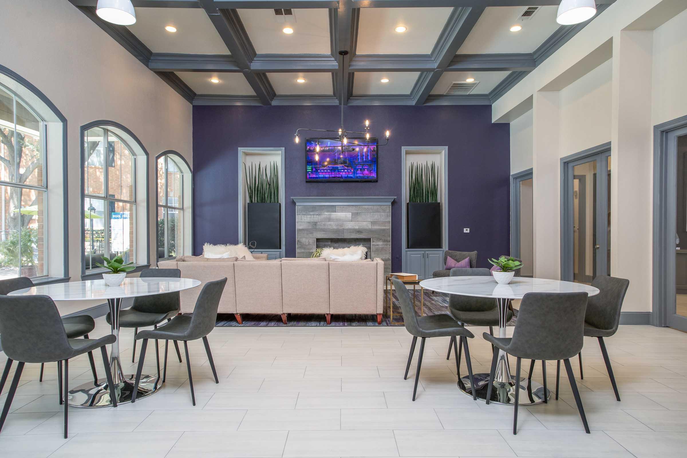 A modern lounge area featuring two round dining tables with black chairs. In the background, a cozy seating area with a light-colored sofa and an electric fireplace is visible. A large television hangs on the wall, surrounded by decorative plants and soft lighting from the coffered ceiling.